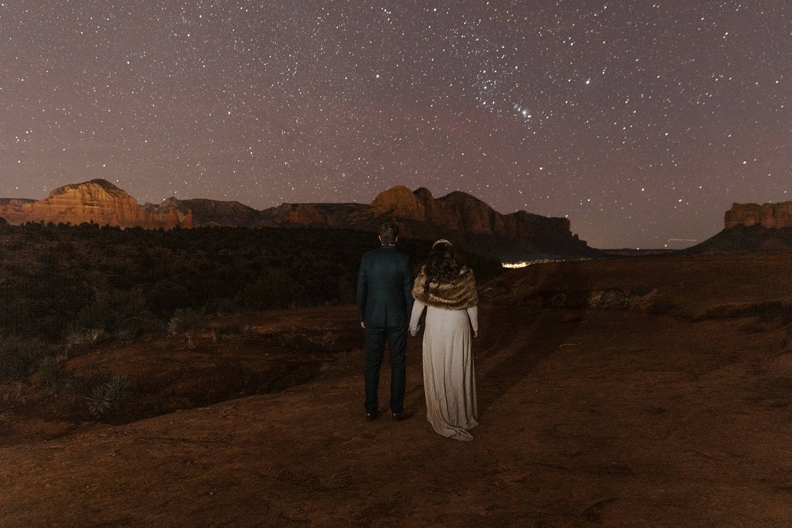 A bride and groom stand hand-in-hand under a star-filled night sky, facing the red rock formations of Sedona, Arizona. The bride wears a long white dress with a fur shawl, and the groom is in a dark suit. The landscape is softly illuminated, highlighting the desert terrain and distant cliffs, creating a romantic and serene atmosphere.