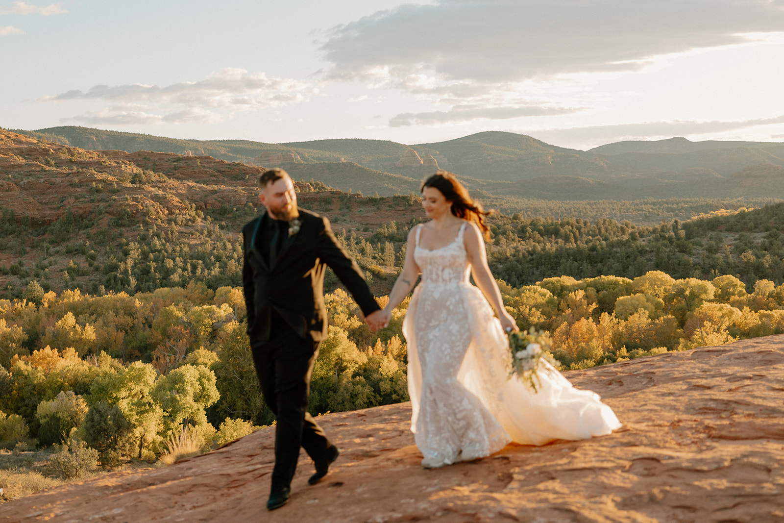 Couple walking hand-in-hand on red rocks, bride in shimmering lace elopement dress.