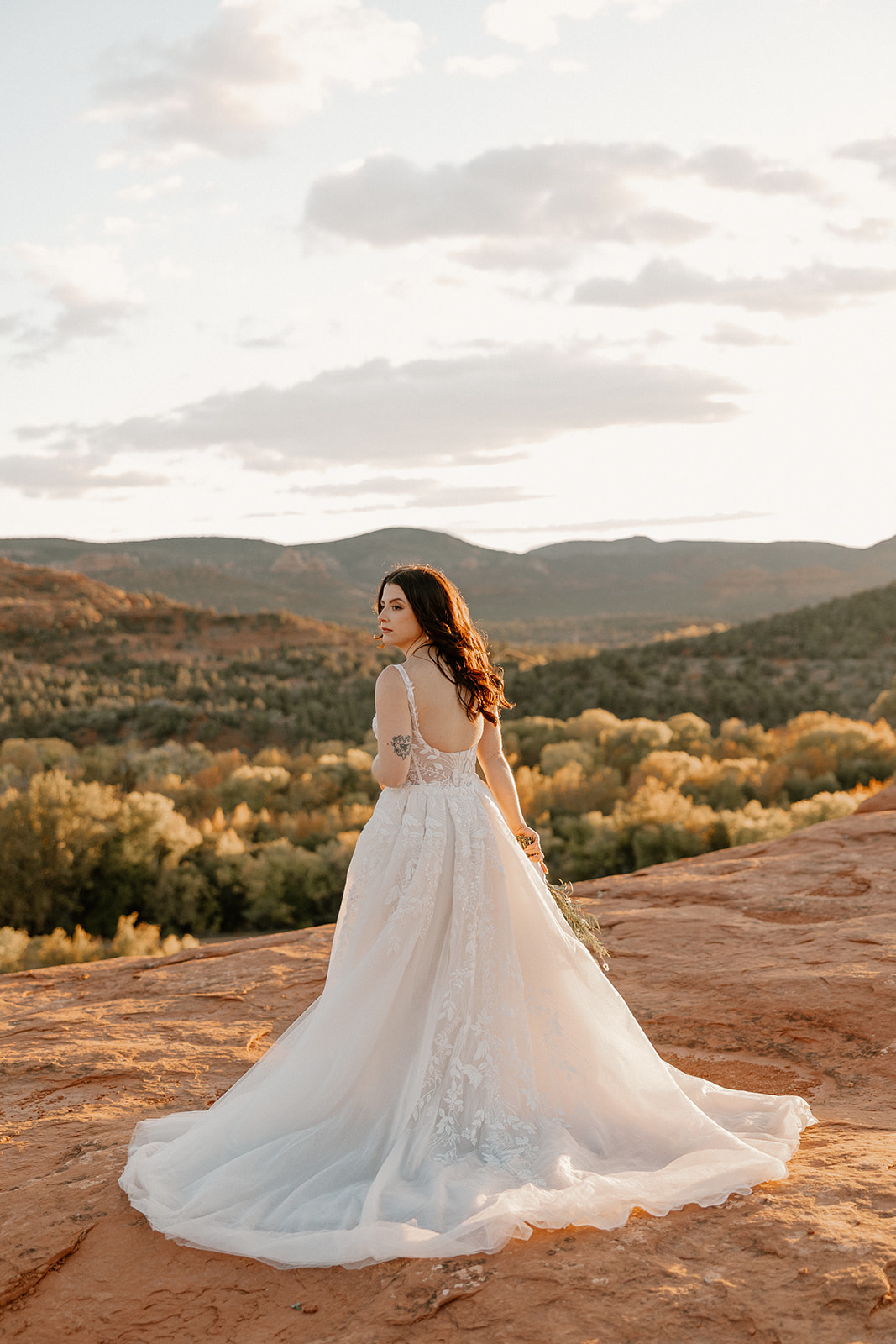 Bride in flowing lace elopement dress looking over shoulder with Sedona landscape behind.