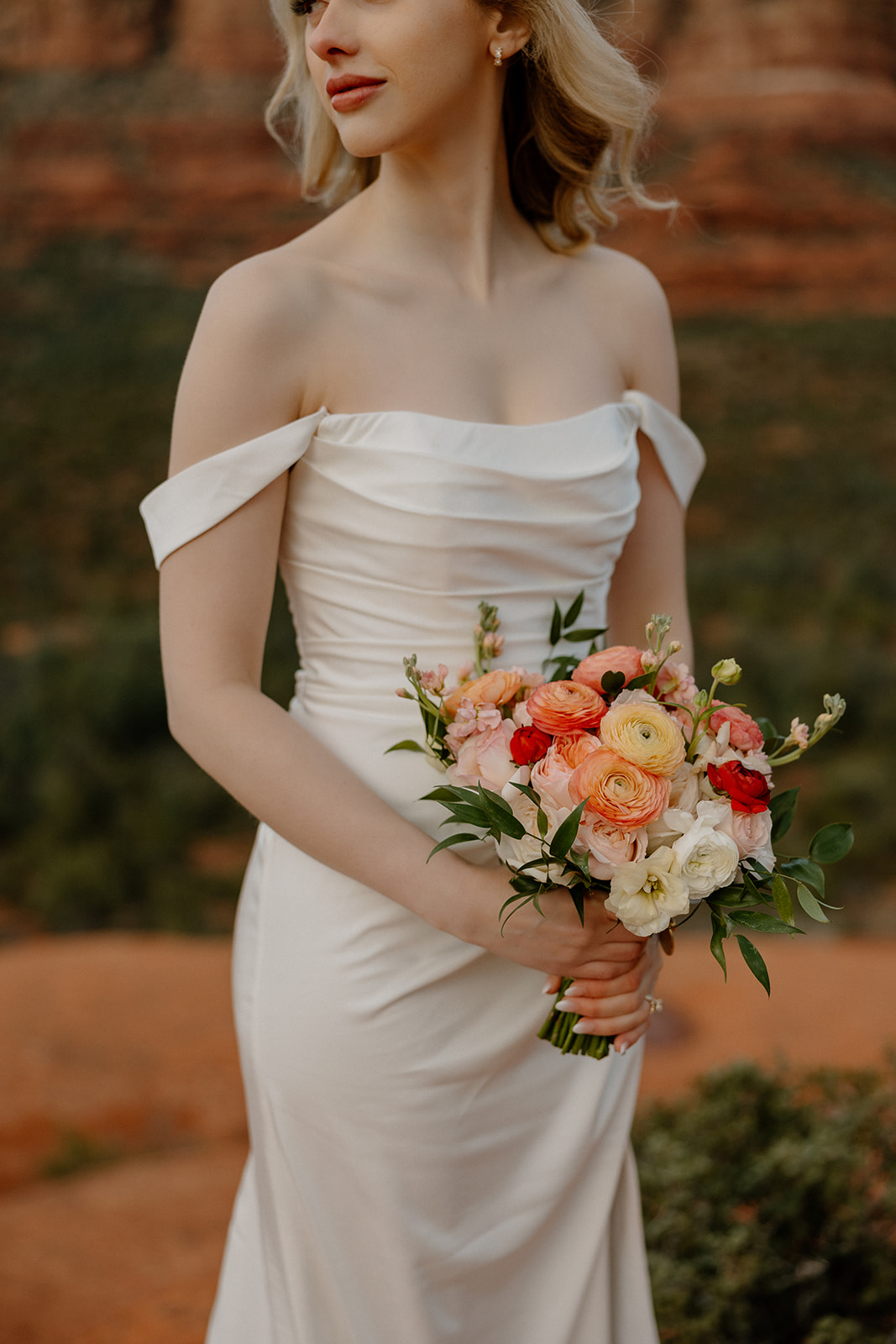 Bride in off-the-shoulder satin elopement dress holding vibrant bouquet in the desert.
