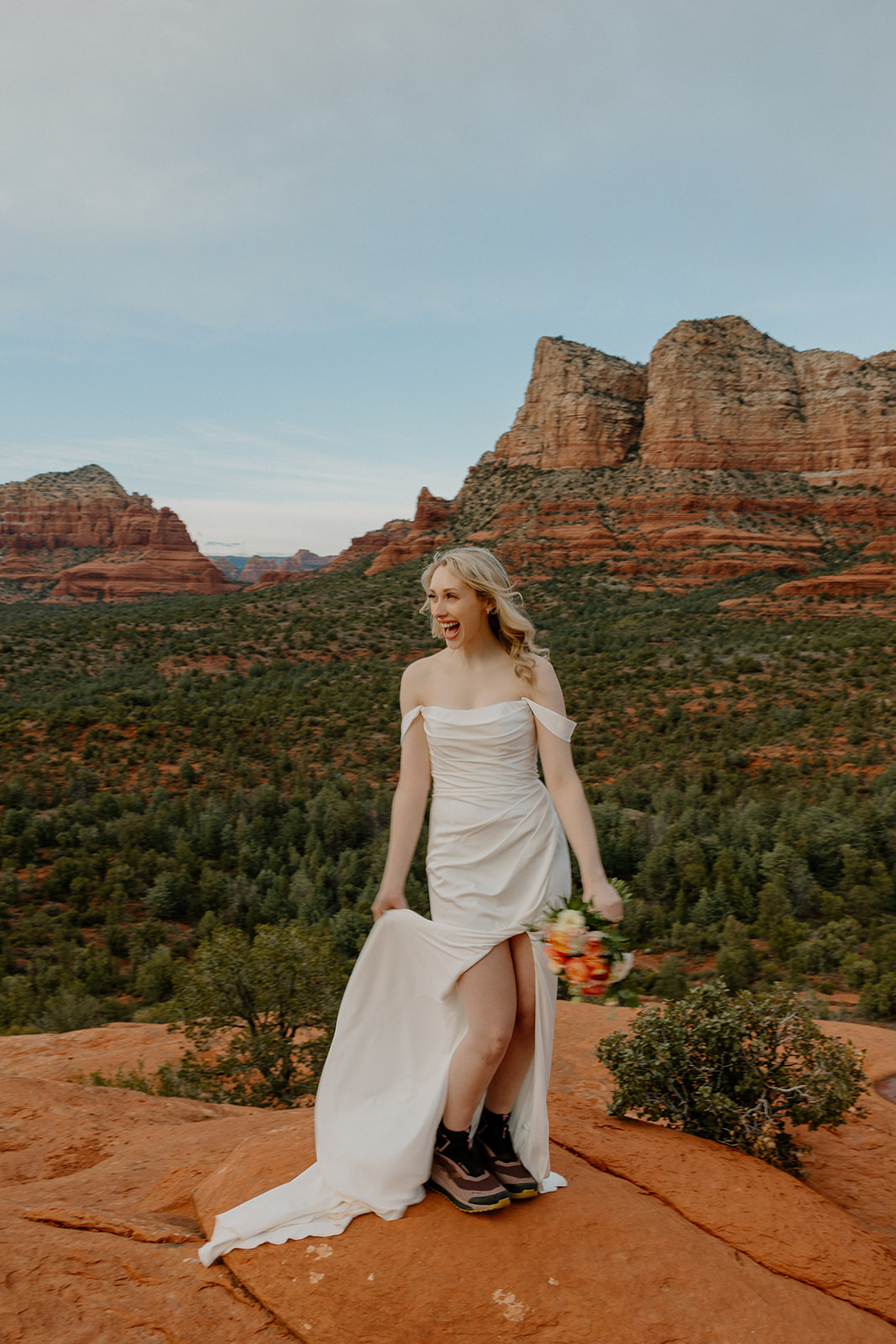 Bride hiking in Sedona wearing off-the-shoulder elopement dress and boots, holding bouquet.