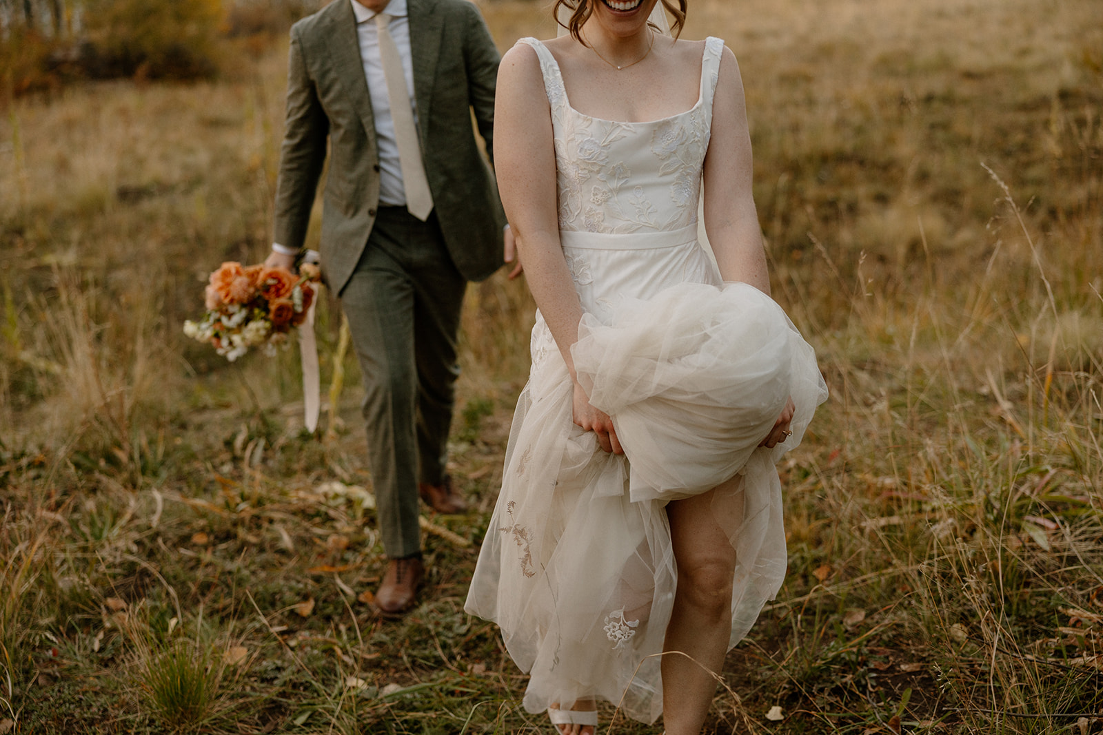 Bride lifting hem of her elopement dress while walking through grassy meadow.