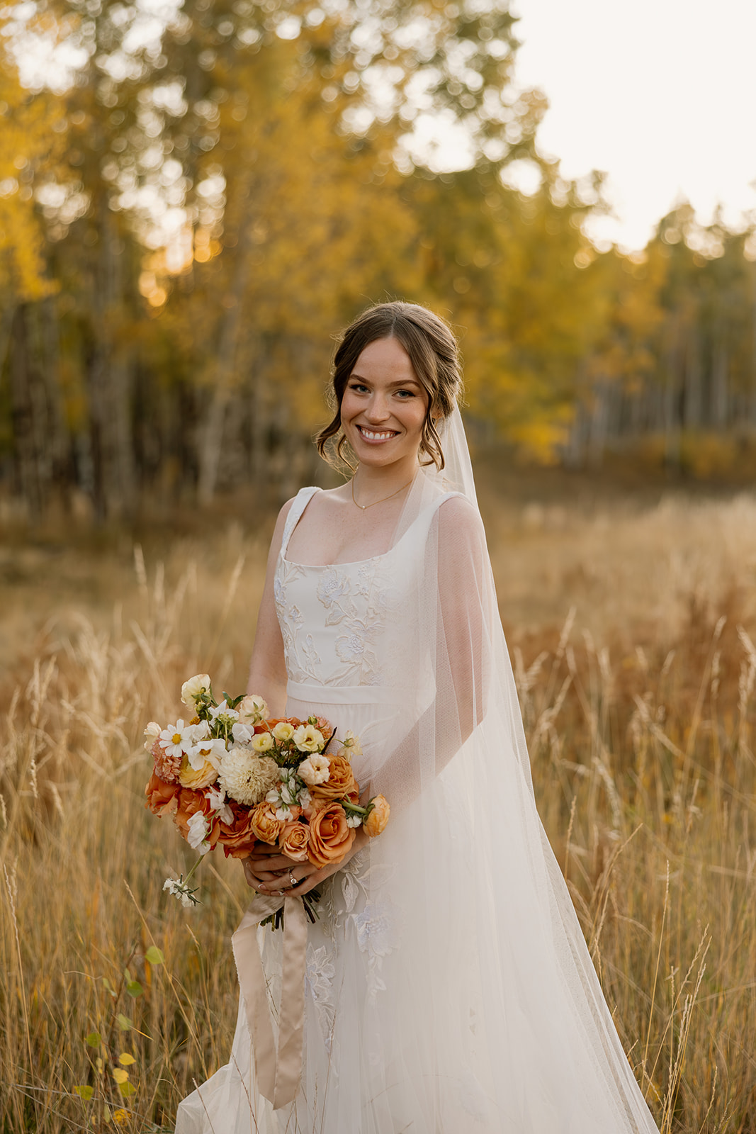 Bride smiling in autumn field, holding orange and cream bouquet, wearing lace elopement dress with veil.