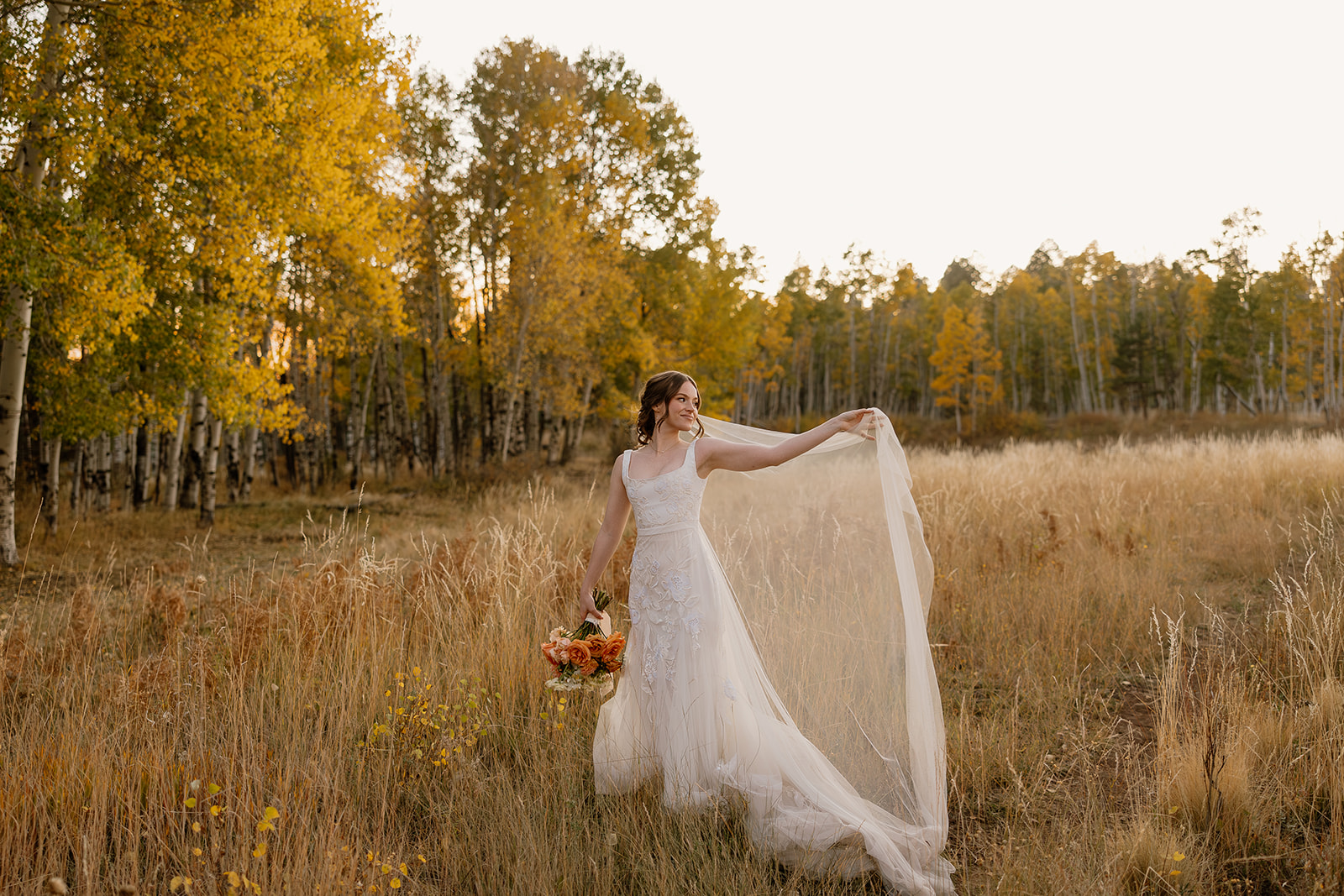 Bride in lace elopement dress holding bouquet and veil in a golden autumn meadow.
