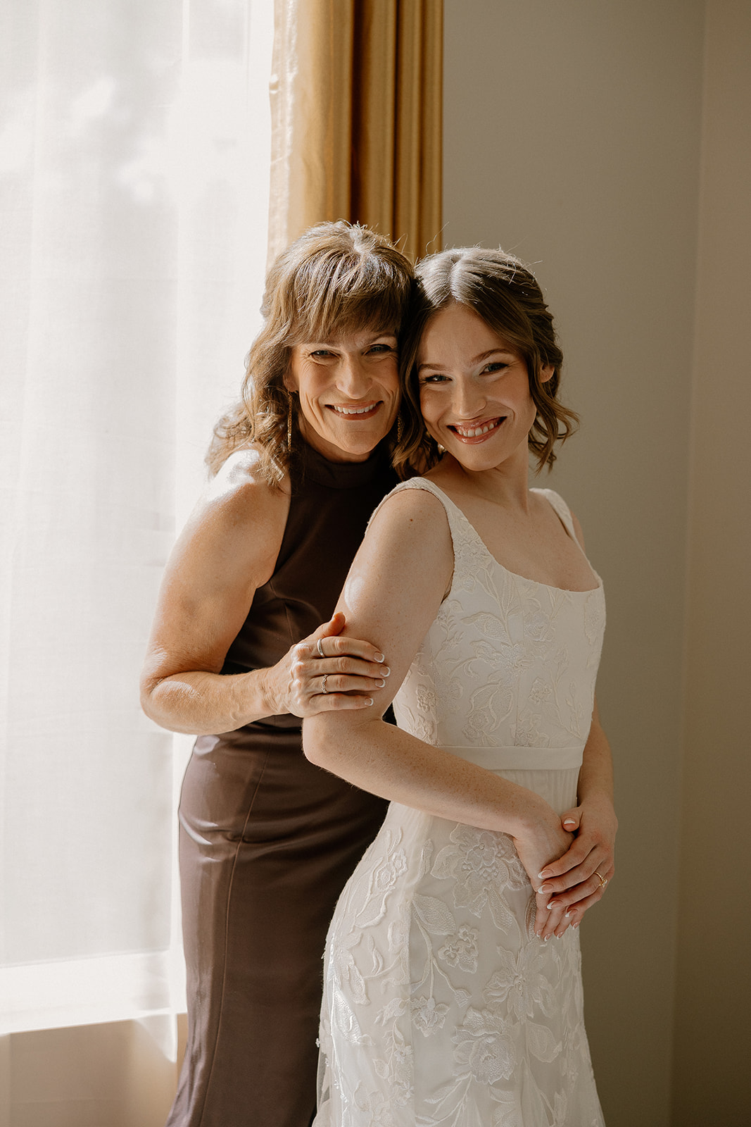 Bride in embroidered elopement dress posing with her mother by a sunlit window.