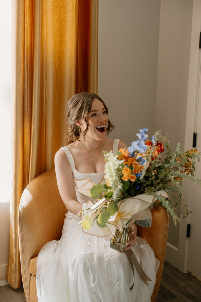 Bride in tulle elopement dress smiling with colorful bouquet while sitting in a cozy chair.