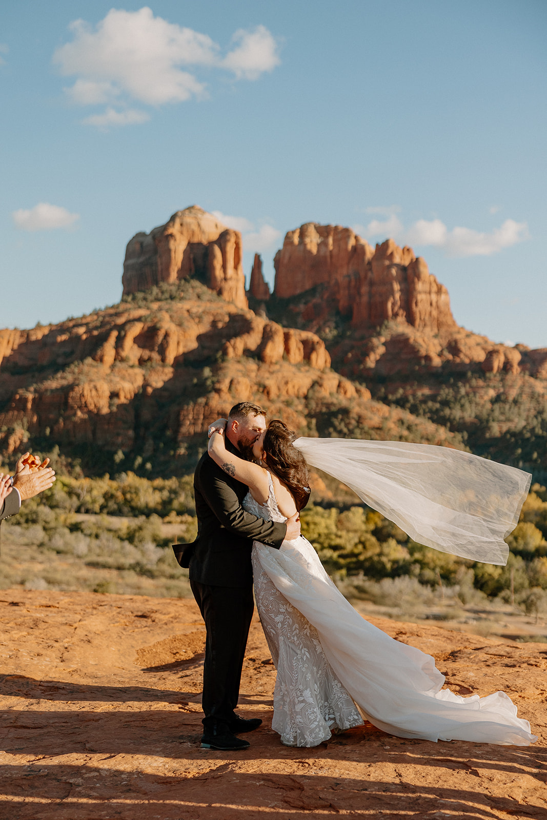 Couple kissing during Sedona ceremony, bride’s elopement dress train and veil blowing in the wind.