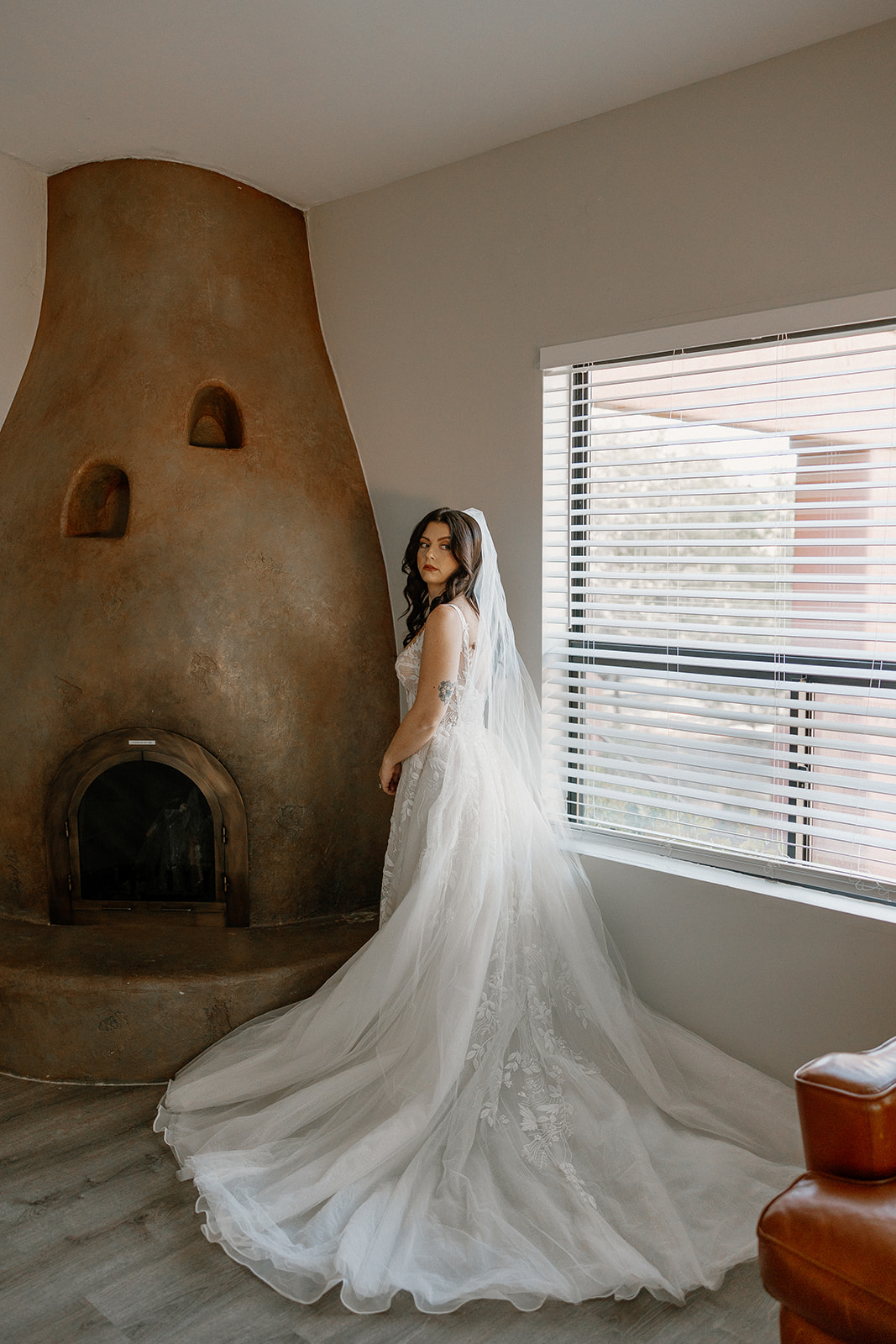 Bride in fitted lace elopement dress with long train and veil, standing by adobe fireplace indoors.