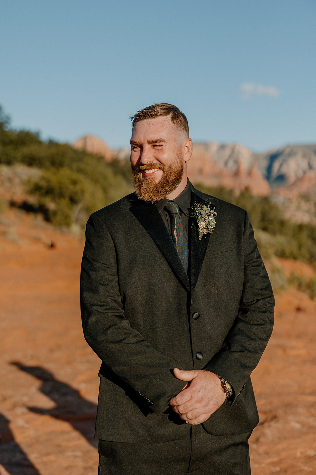 Groom wedding attire in a matte black suit, smiling confidently under the Arizona sun with Sedona’s red rocks in the background.