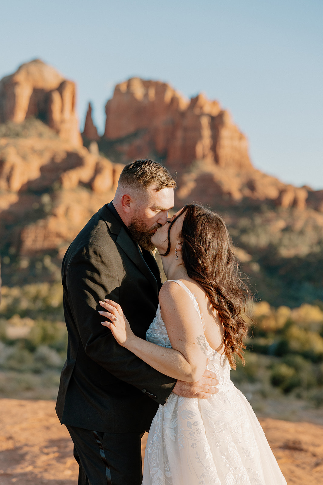 Groom wedding attire styled in a classic black suit as the groom kisses his bride against a golden hour desert backdrop.