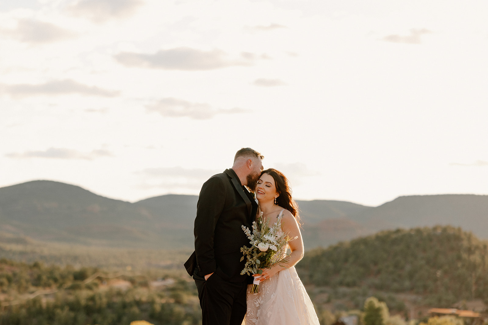 Groom wedding attire in a black suit and tie as he kisses his bride’s temple during sunset, with a soft bouquet and desert skyline.