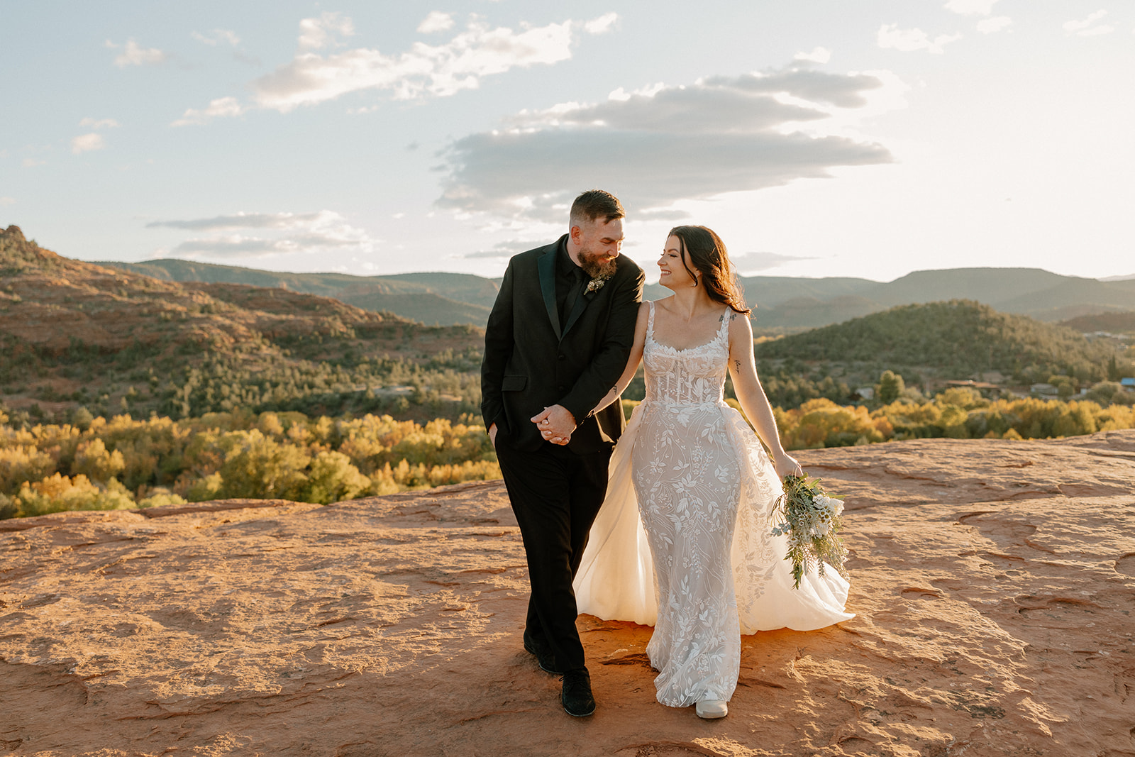 Groom wedding attire styled in a modern black suit as the couple walks hand-in-hand across desert red rock during sunset.