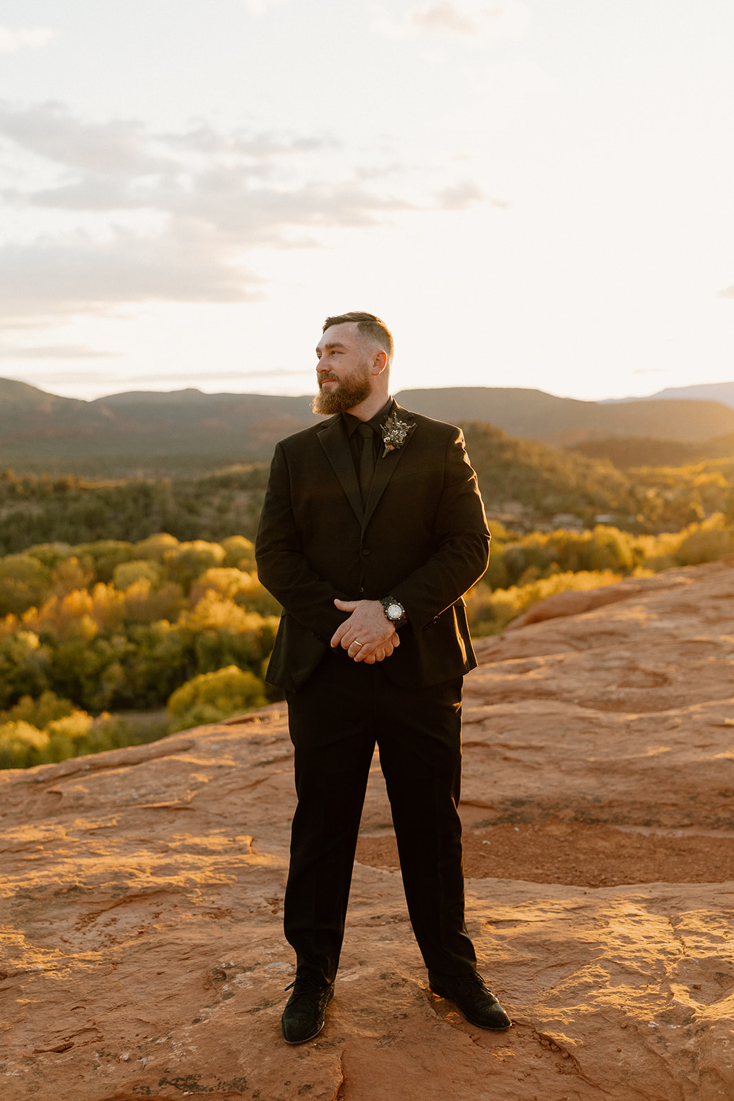 Editorial-style groom wedding attire featuring a sleek black suit, captured solo at golden hour on red rock terrain.