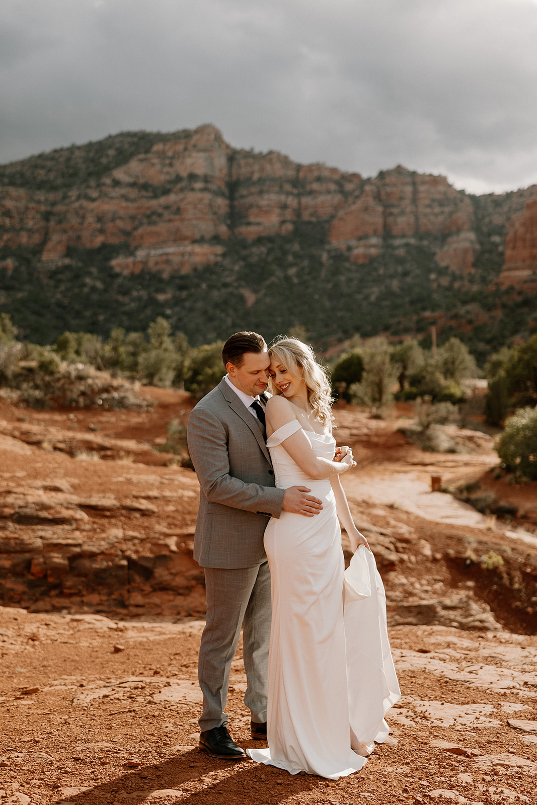 Groom wedding attire in a soft gray suit with a black tie, embracing his bride on red rock terrain beneath dramatic desert skies in Sedona.