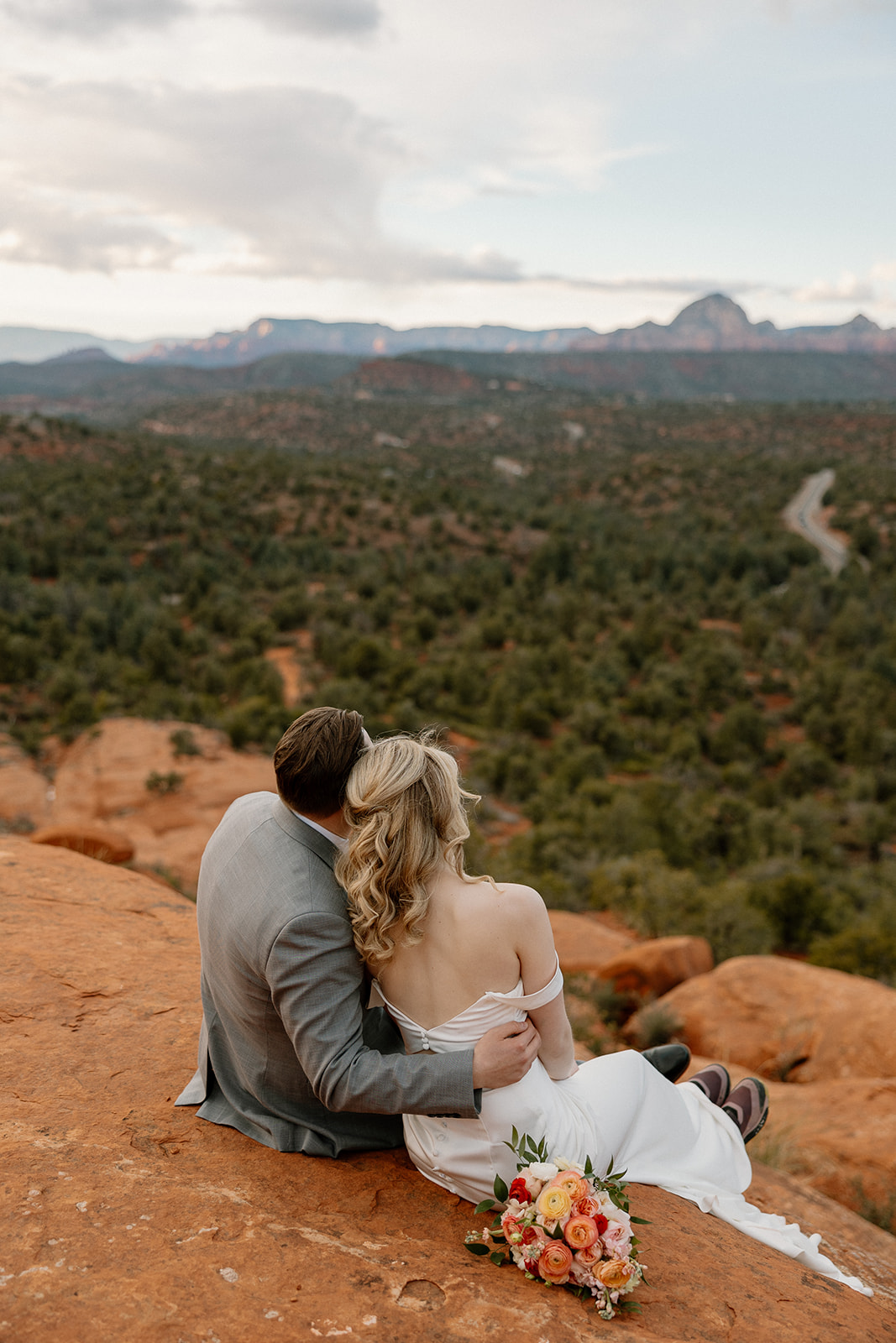 A groom in a light gray suit embraces his bride on a red rock overlook, showcasing relaxed and romantic groom wedding attire for a scenic Sedona elopement.