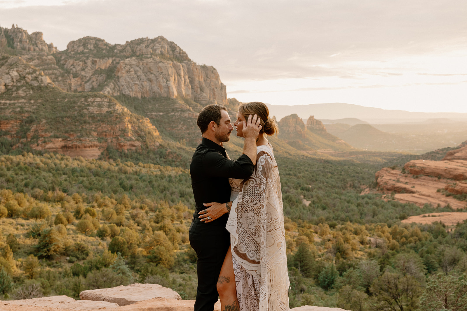 Groom wedding attire in an all-black look, embracing his bride at sunset on a cliff above Sedona’s lush, expansive valley.