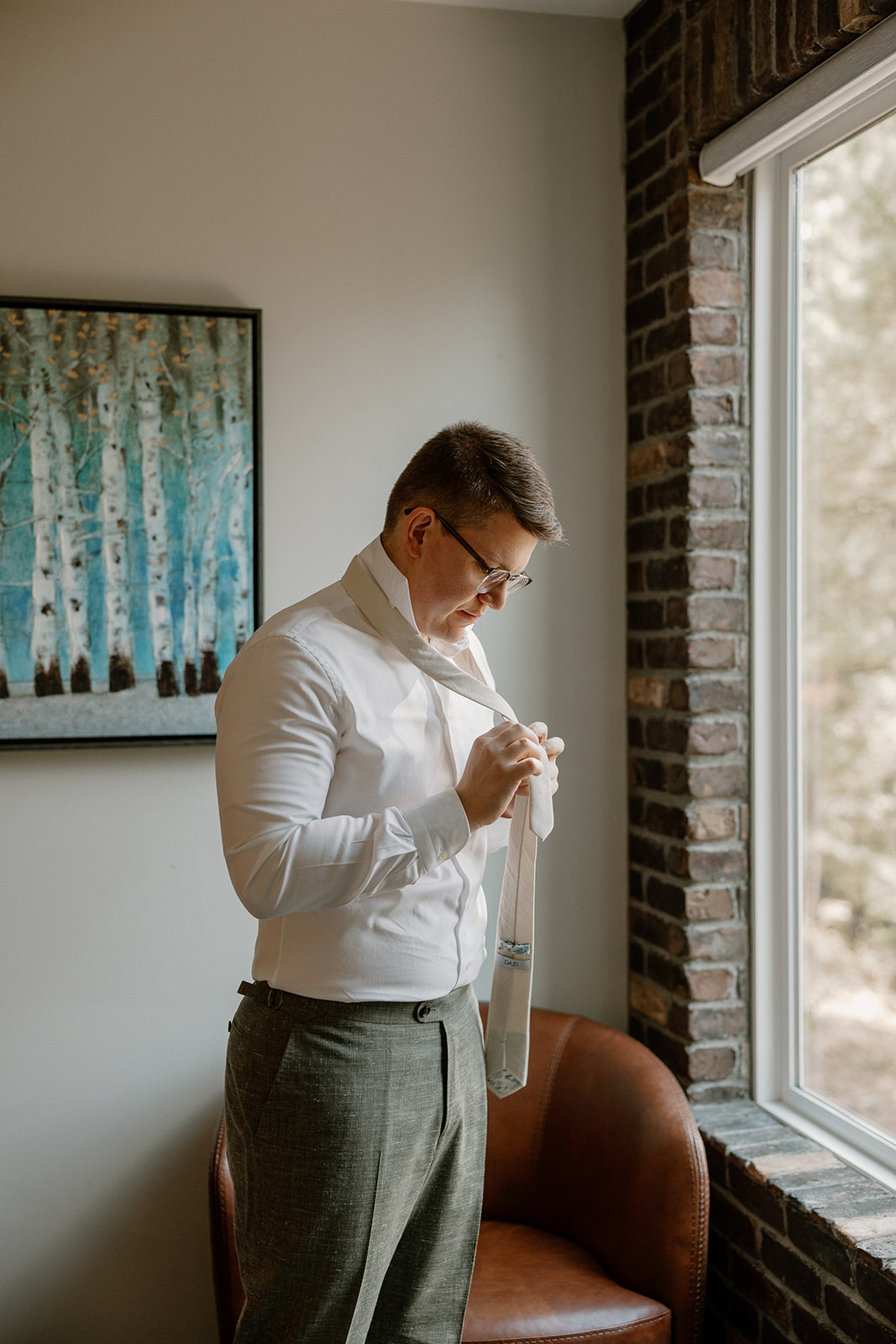 Groom wedding attire prep featuring a crisp white shirt and soft green trousers, tying a neutral-colored tie near a sunlit window.