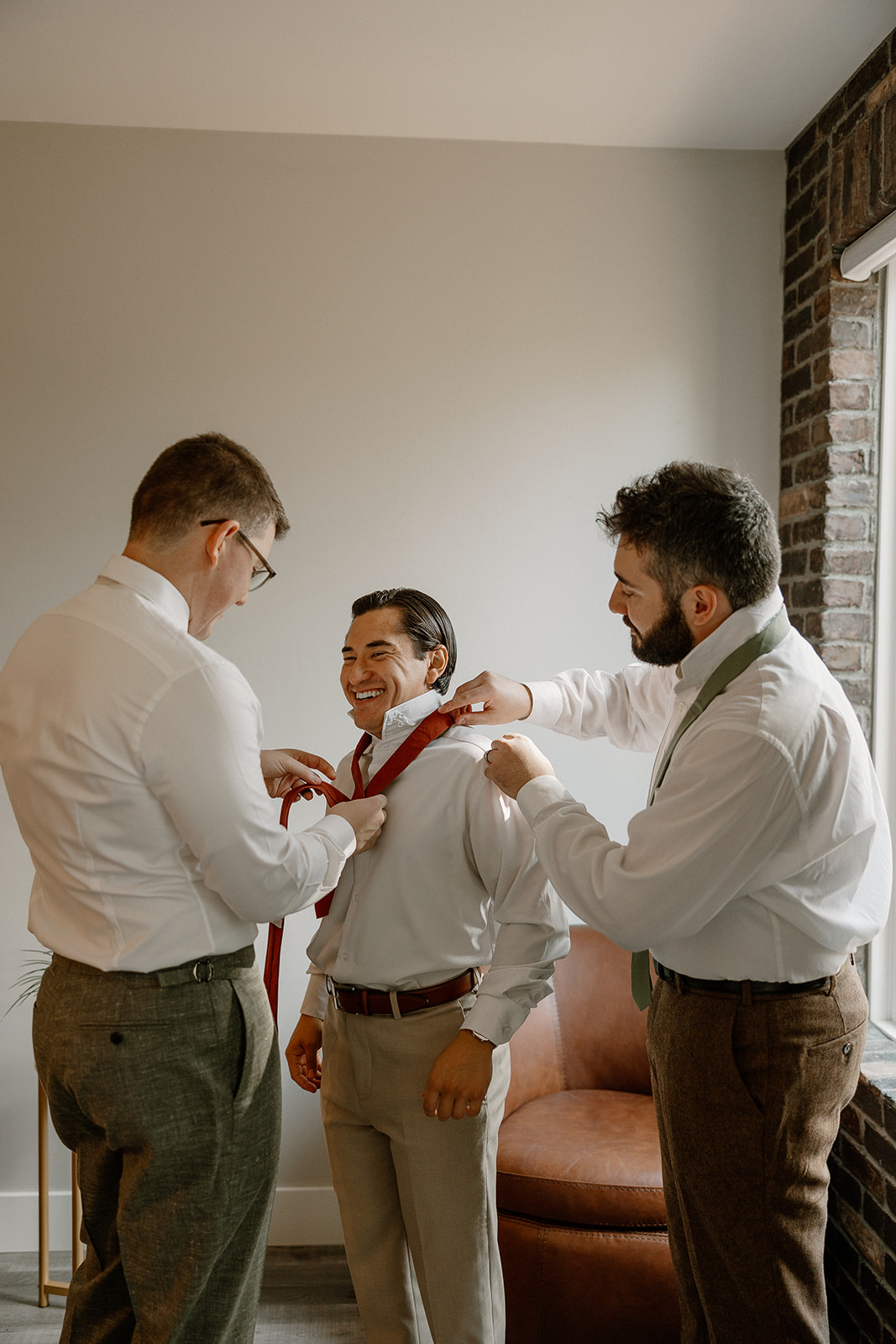 Groom wedding attire prep moment with groomsmen tying a red tie, highlighting earthy tones and laid-back wedding day vibes.
