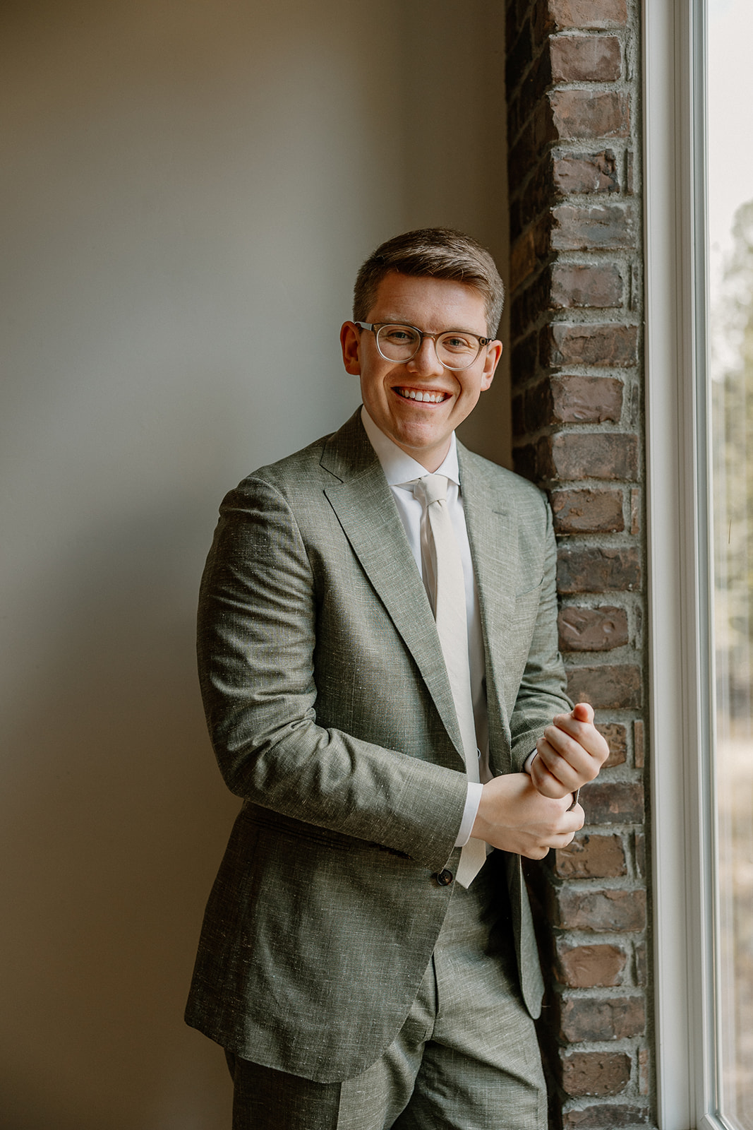 Groom wedding attire in a sage green suit with a neutral tie, smiling by a window just before heading to the ceremony.