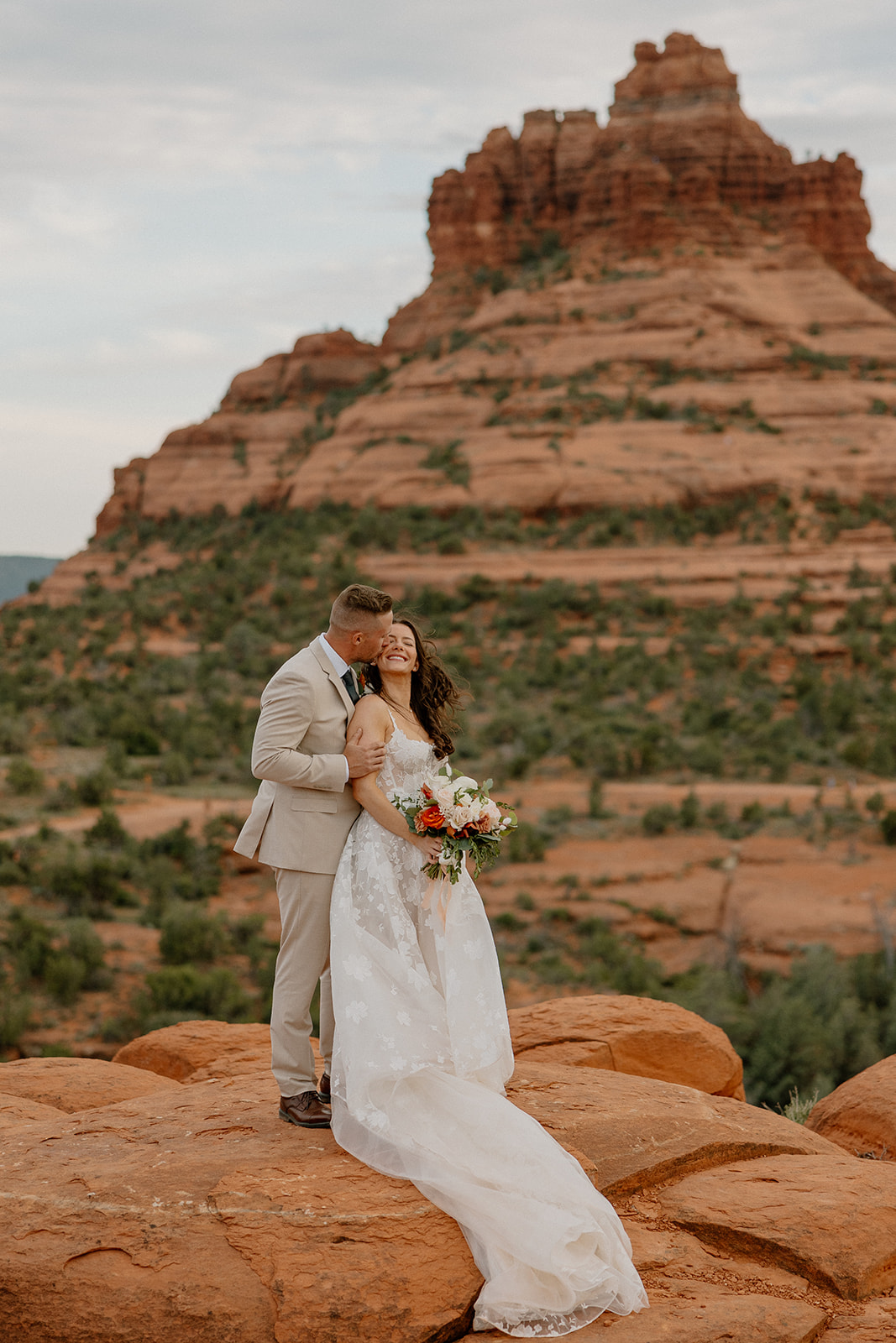 Groom wedding attire in a beige suit paired with brown shoes, holding his bride close in front of Bell Rock’s iconic red sandstone.