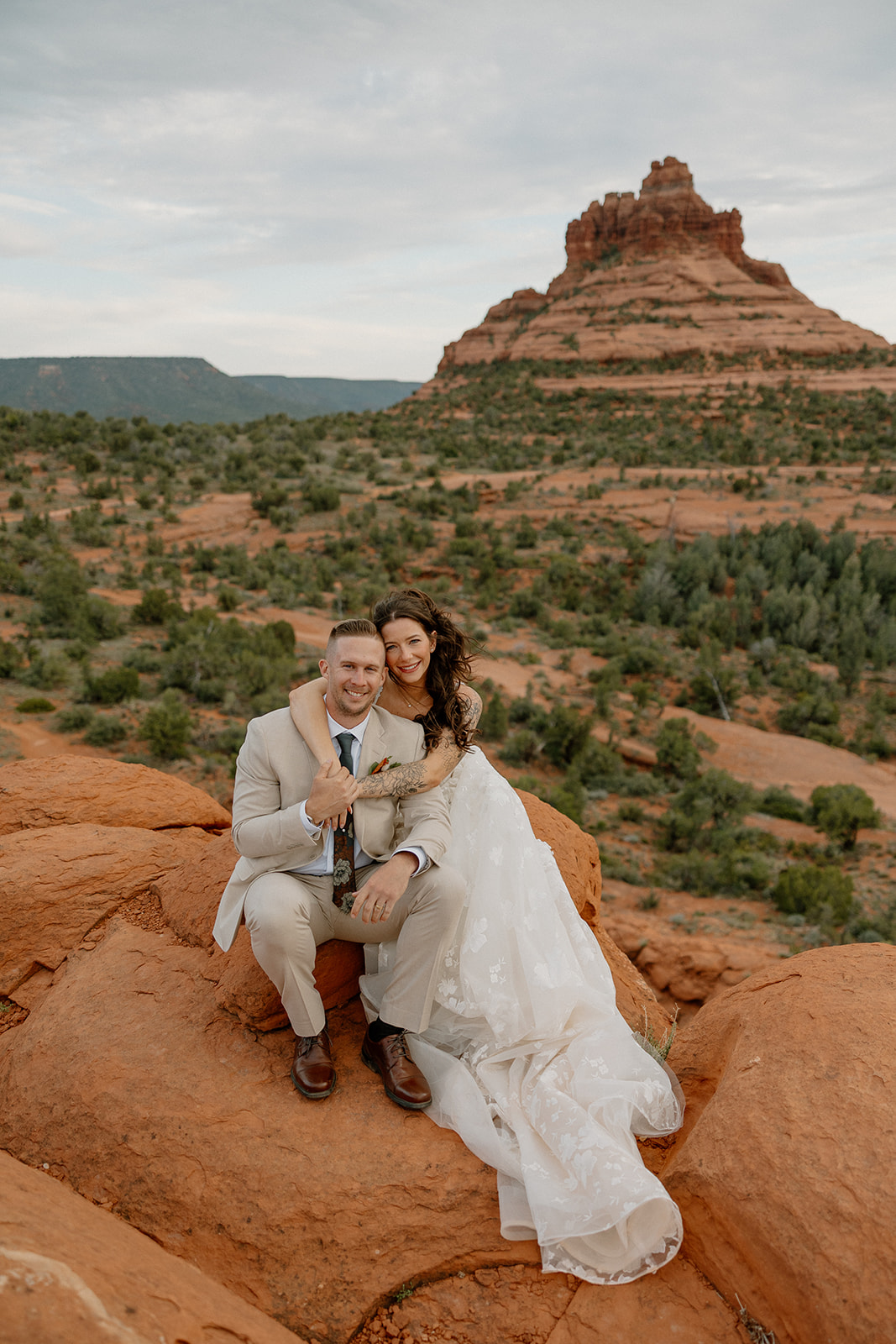 A groom in beige wedding attire sits with his bride on a red rock ledge, his look styled with brown shoes and a muted tie—modern desert groom vibes.