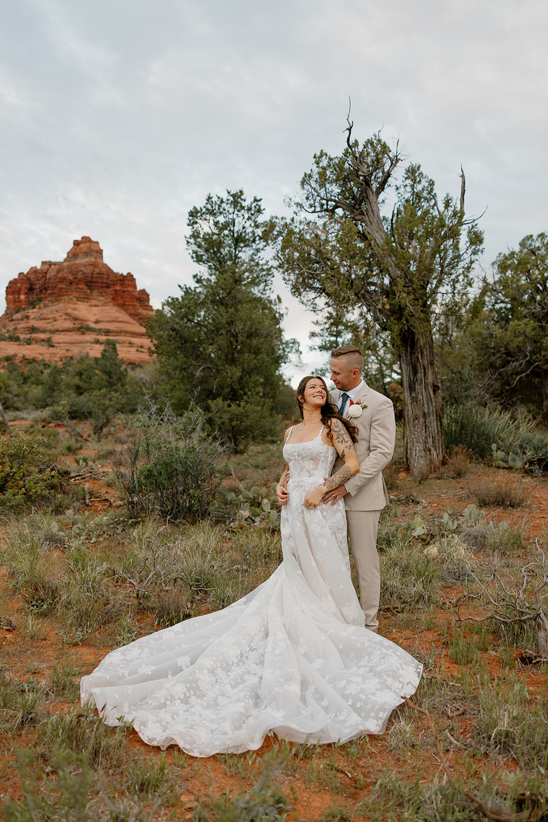 Romantic portrait of groom wedding attire in a beige suit, embracing his bride beneath Sedona pines with Bell Rock in the distance.