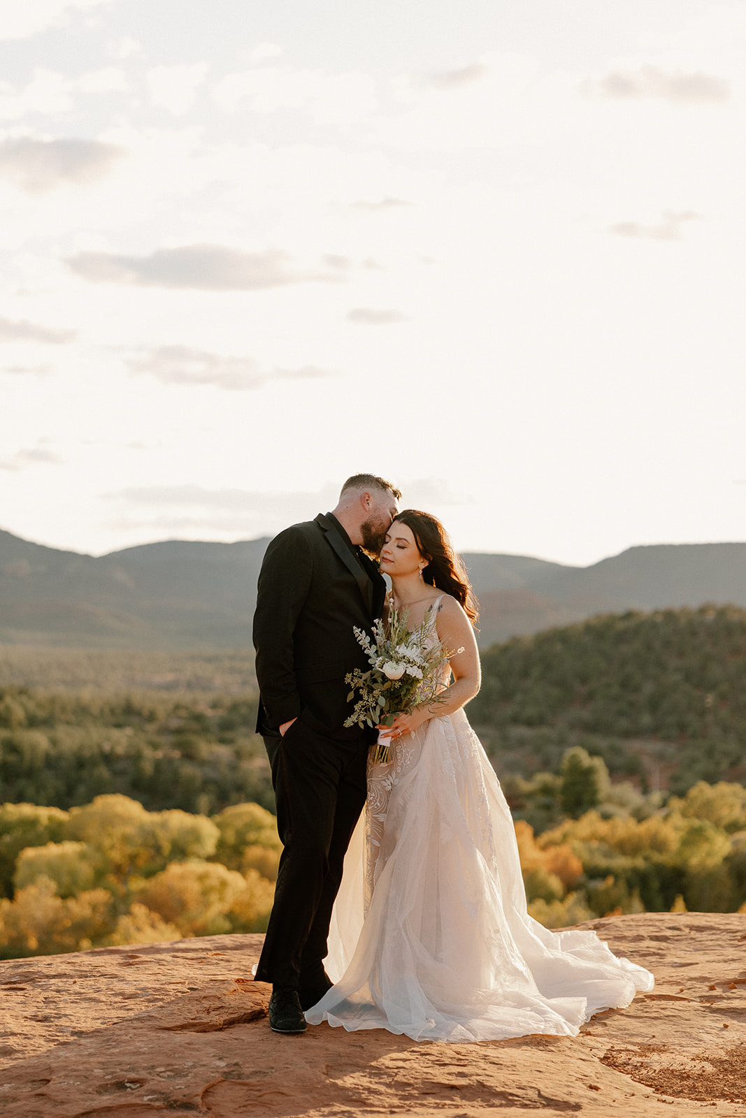 Groom kissing bride’s temple in Sedona, bride in flowing lace elopement dress.