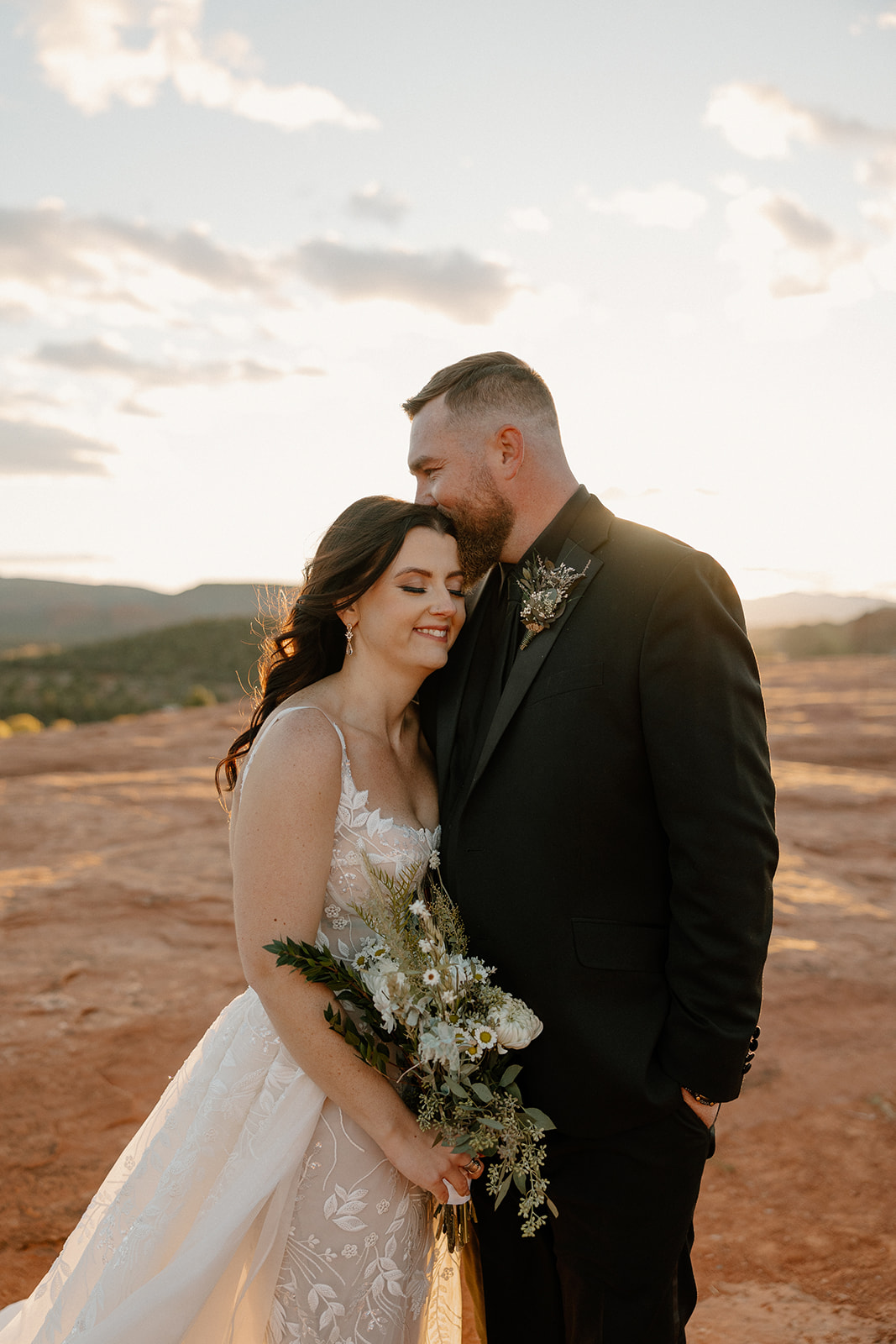 Bride and groom embrace with bride resting head on groom's chest while he kisses her forehead at sunset during outdoor bridals