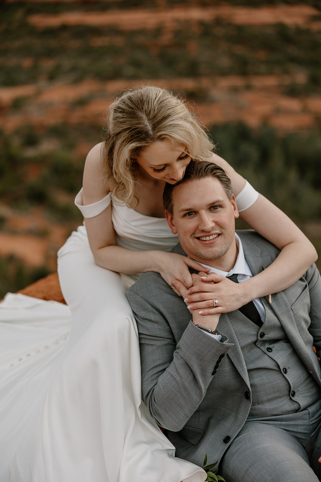 Bride leans over groom with a soft smile, her arms gently wrapped around him as they soak in the quiet red rock views.