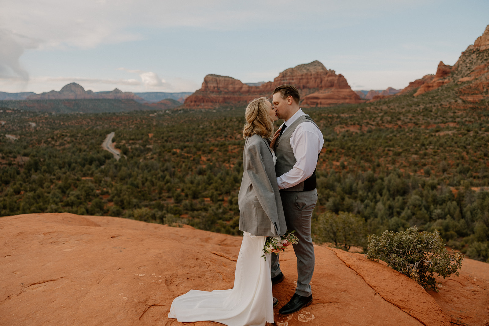 Bride wearing groom's gray suit jacket leans into to kiss her groom on top of red rock area