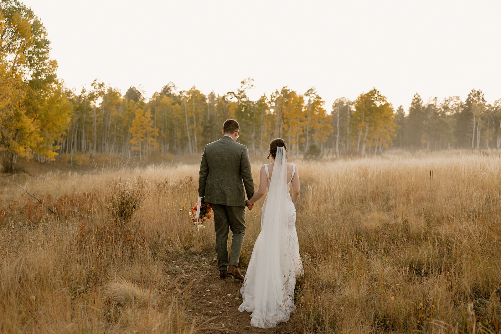 Bride and groom walk through field of golden grass after forest wedding ceremony