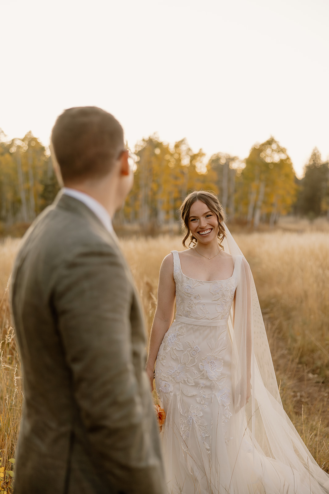 Groom looks over at his glowing bride in front of golden grassy field both joyful after deciding what is the best time to elope
