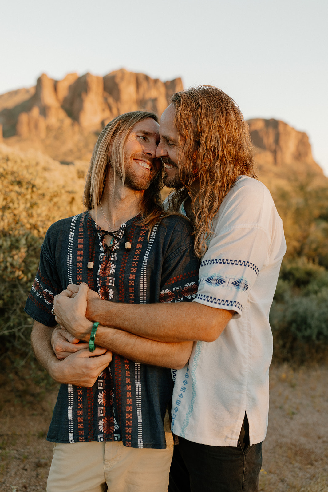 Candid moment between two grooms in casual boho wedding attire, embracing at golden hour in the Arizona desert.