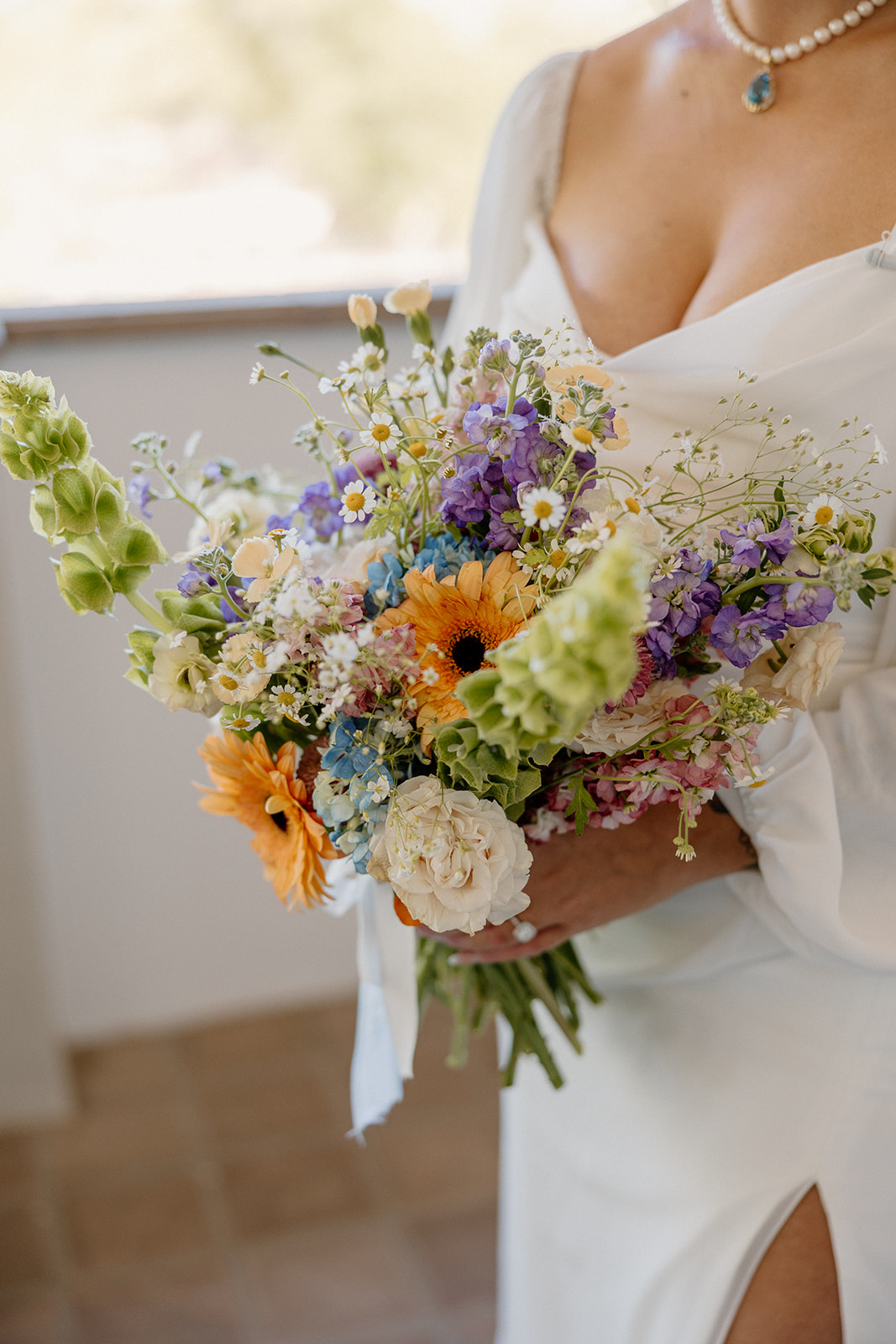 A vibrant bridal bouquet full of wildflowers, daisies, and gerbera blooms, held just beneath the bride’s turquoise pendant necklace.