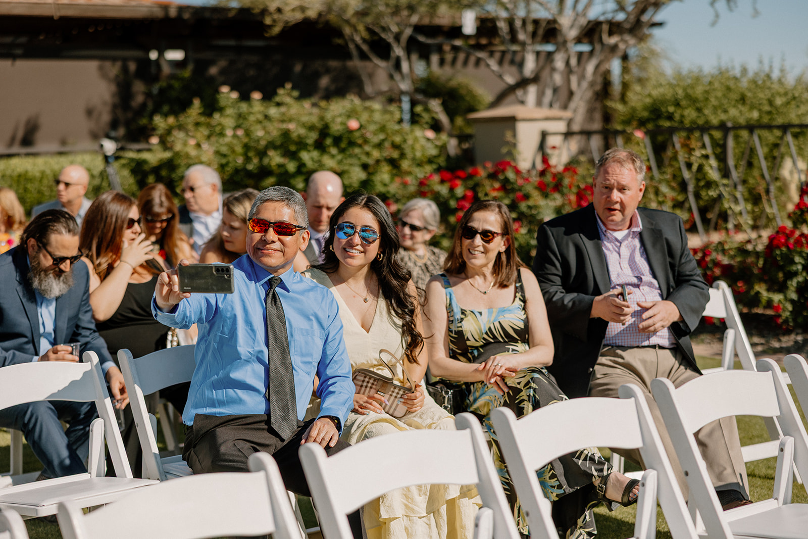 Guests grab selfies and enjoy the sunshine while waiting for the ceremony to start, surrounded by floral landscaping and bright light.