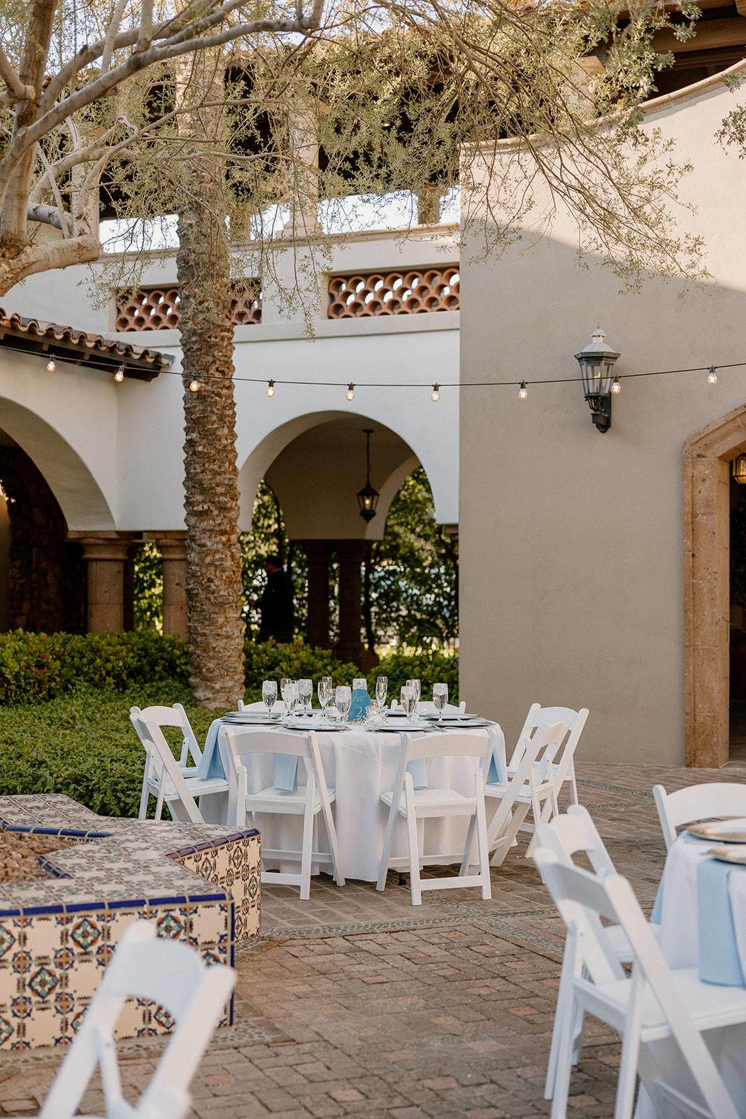 Round tables with white linens set up in the Spanish-style courtyard, shaded by olive trees and twinkle lights at Blackstone Country Club.