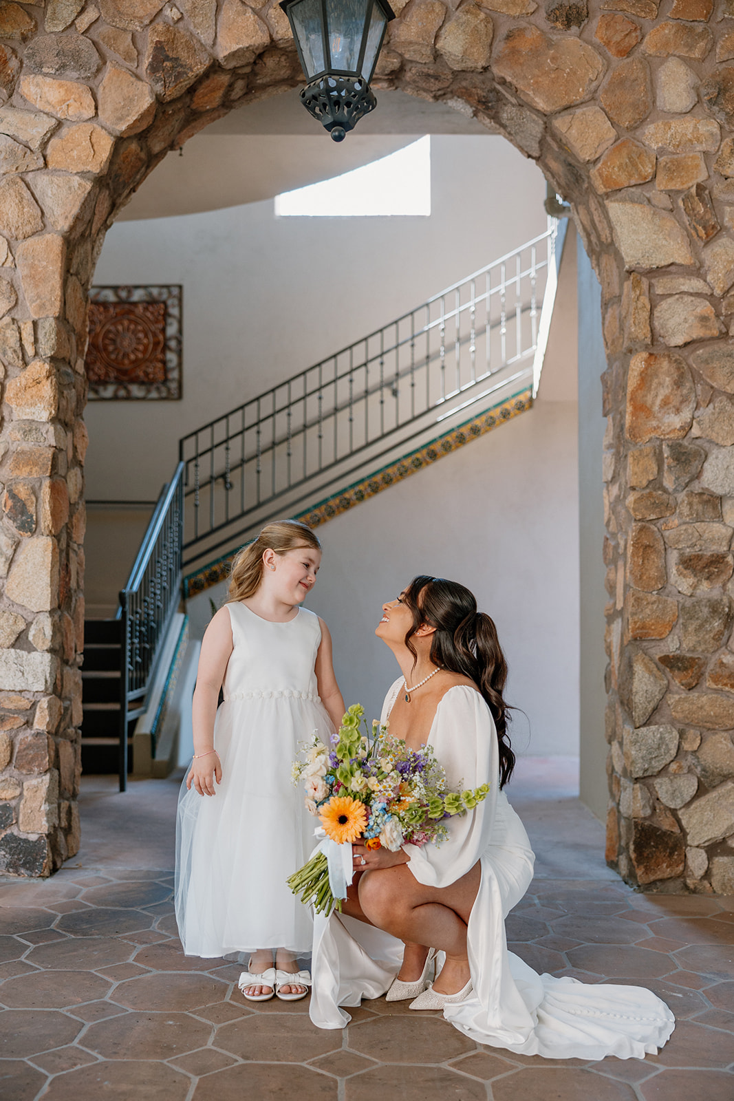 The bride kneels to chat with the flower girl under a stone archway at Blackstone Country Club, both glowing with joy.