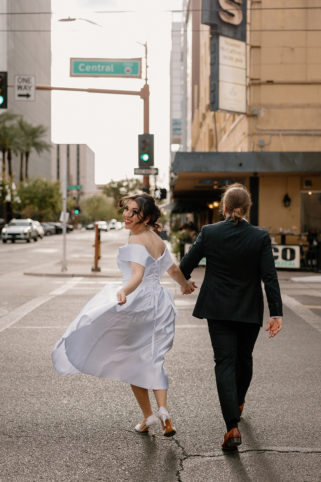 Bride playfully looking over her shoulder while holding hands and running with her partner across Central Avenue.