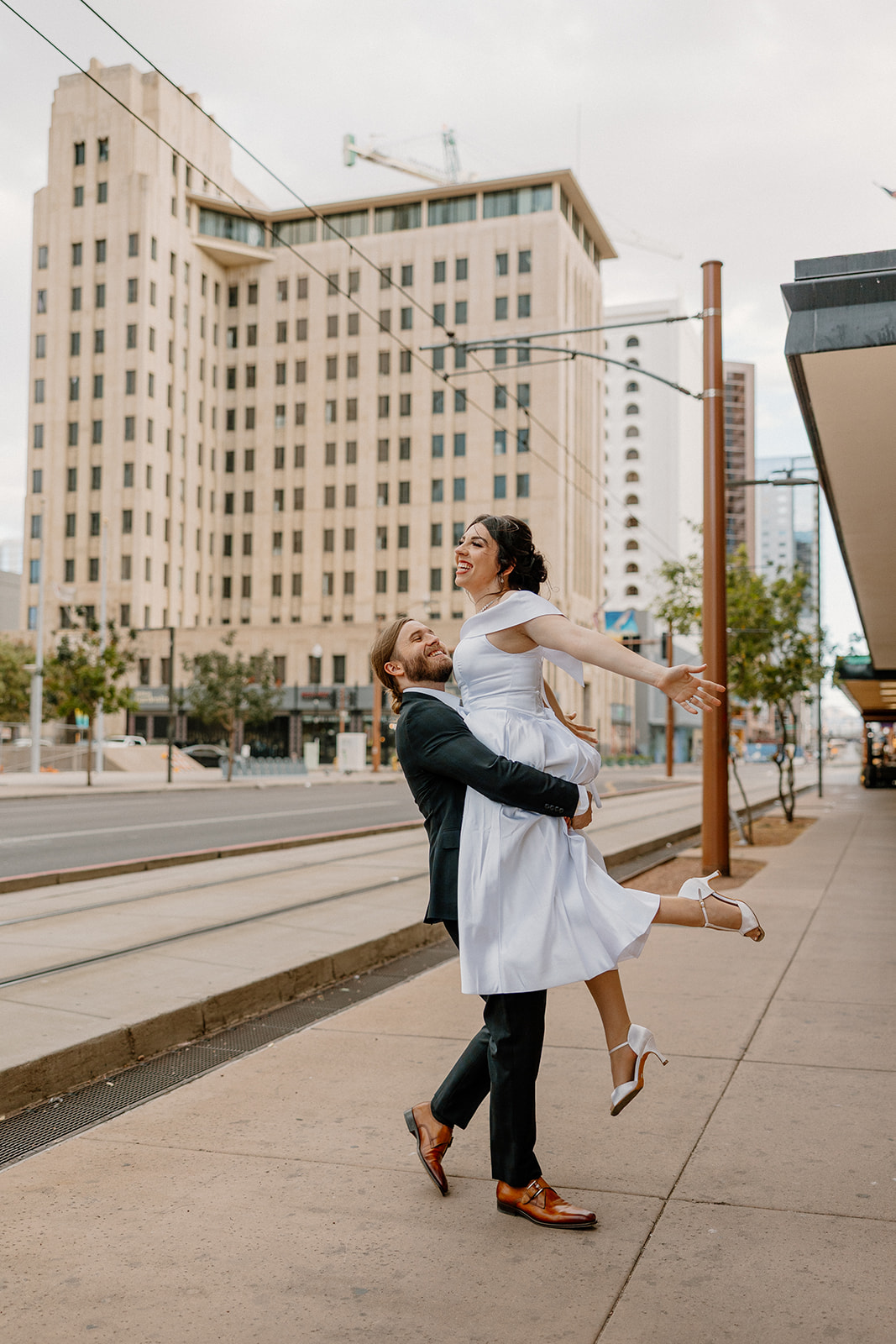 Man lifting woman in a white dress on a city sidewalk, surrounded by downtown Phoenix buildings.