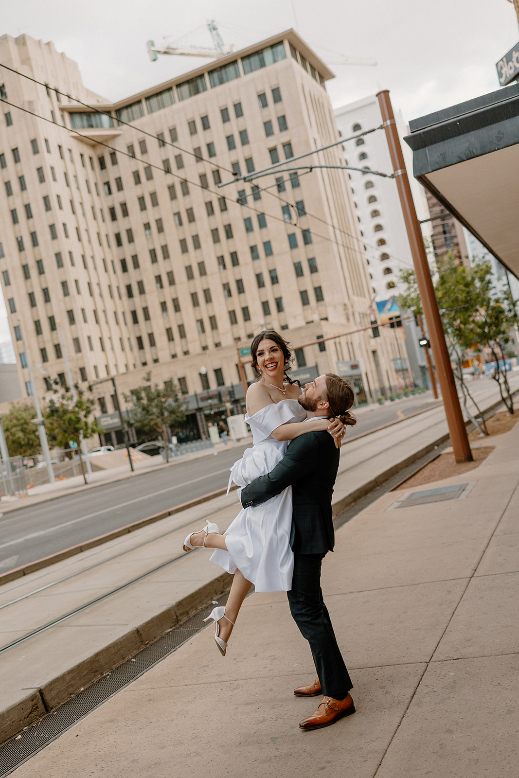 Man lifting his fiancée on the sidewalk, both smiling with tall buildings in the background.