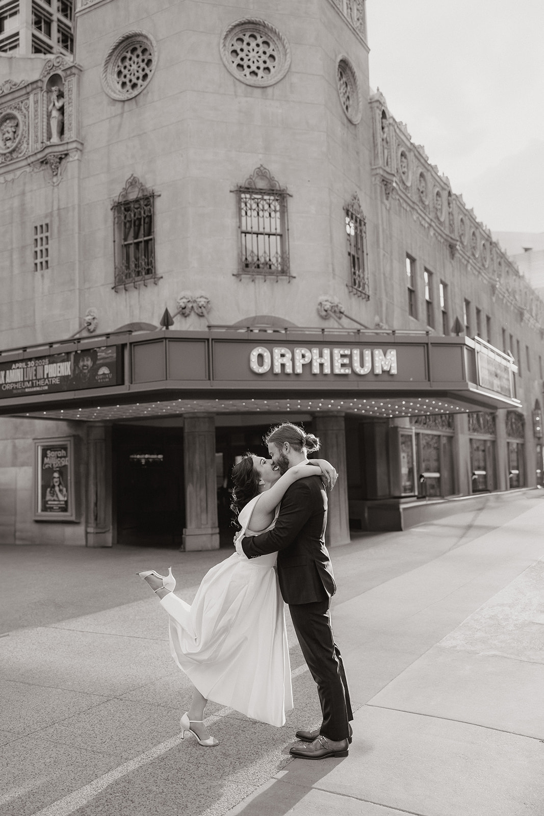 Bride being lifted in a joyful embrace outside the Orpheum Theatre in downtown Phoenix, captured in black and white during their engagement session.