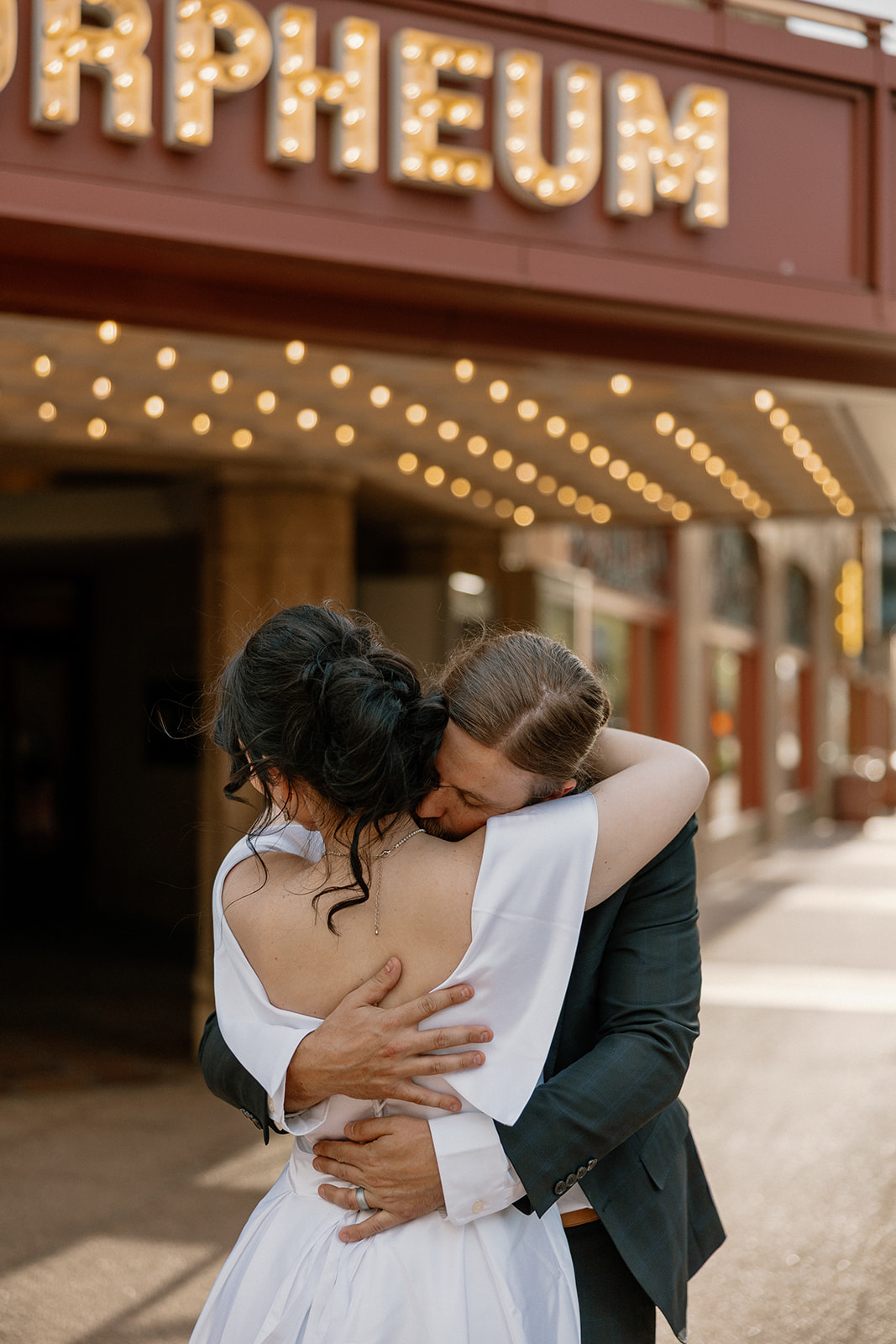 Groom hugging bride from behind under the glowing Orpheum Theatre sign, mid-embrace.