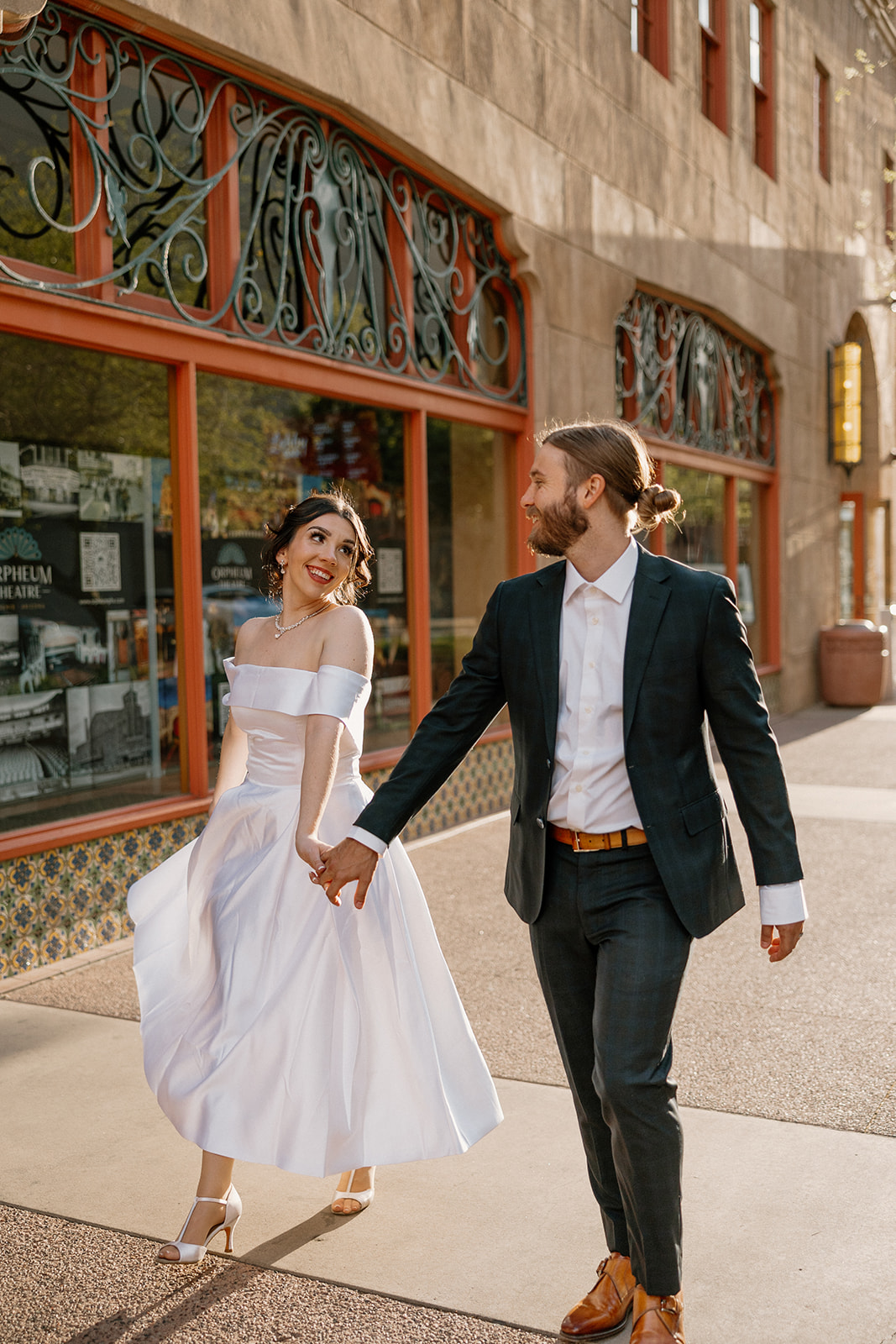 Bride smiling at her partner while walking hand-in-hand outside a historic downtown theater.