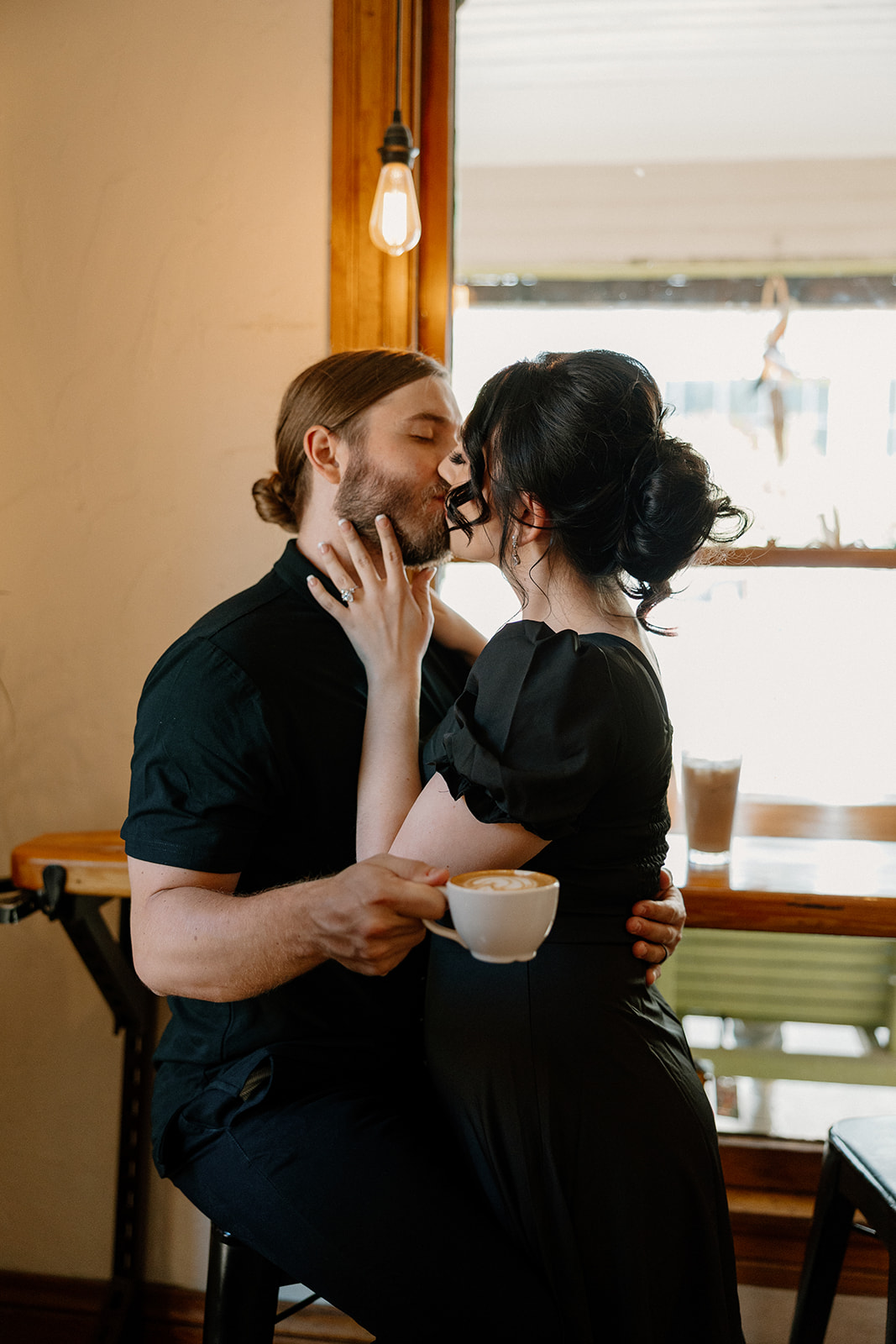 Couple kissing while sitting on a barstool, with a cappuccino in hand and coffee shop lighting in the background.