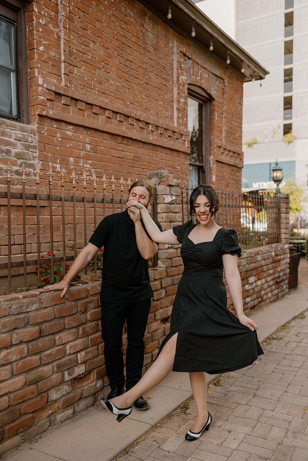 Groom gently kissing bride’s hand as she smiles and twirls beside a red brick building.