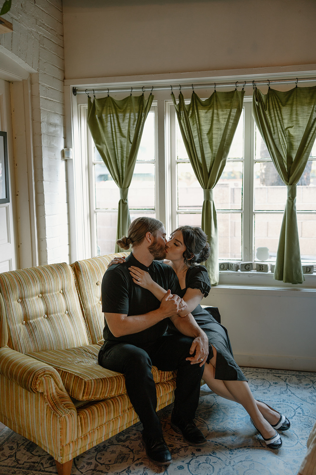 Couple kissing on a yellow retro couch in front of a window with green curtains during their coffee shop engagement session.