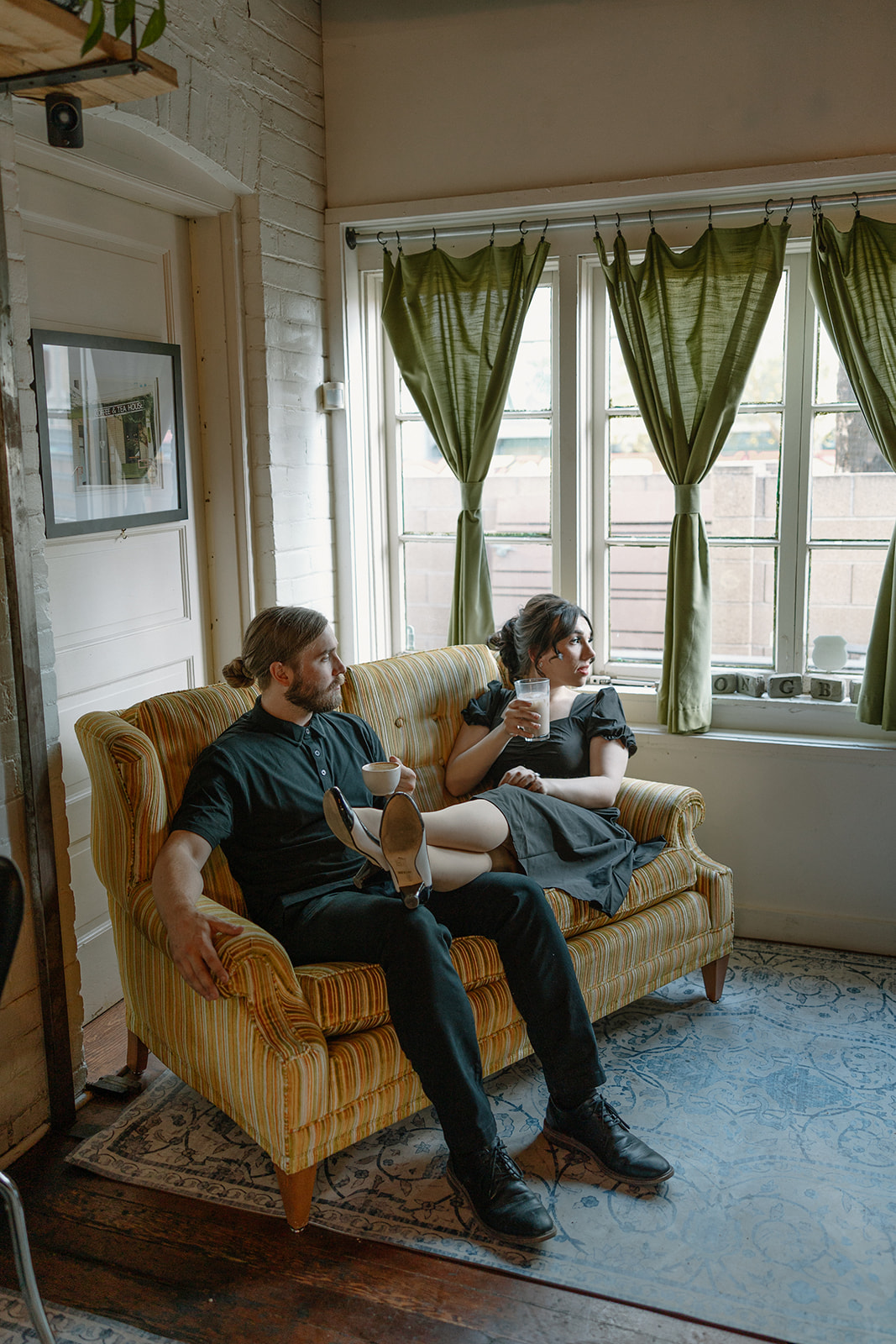 Couple lounging with drinks in hand on a vintage couch during their coffee shop engagement photos.