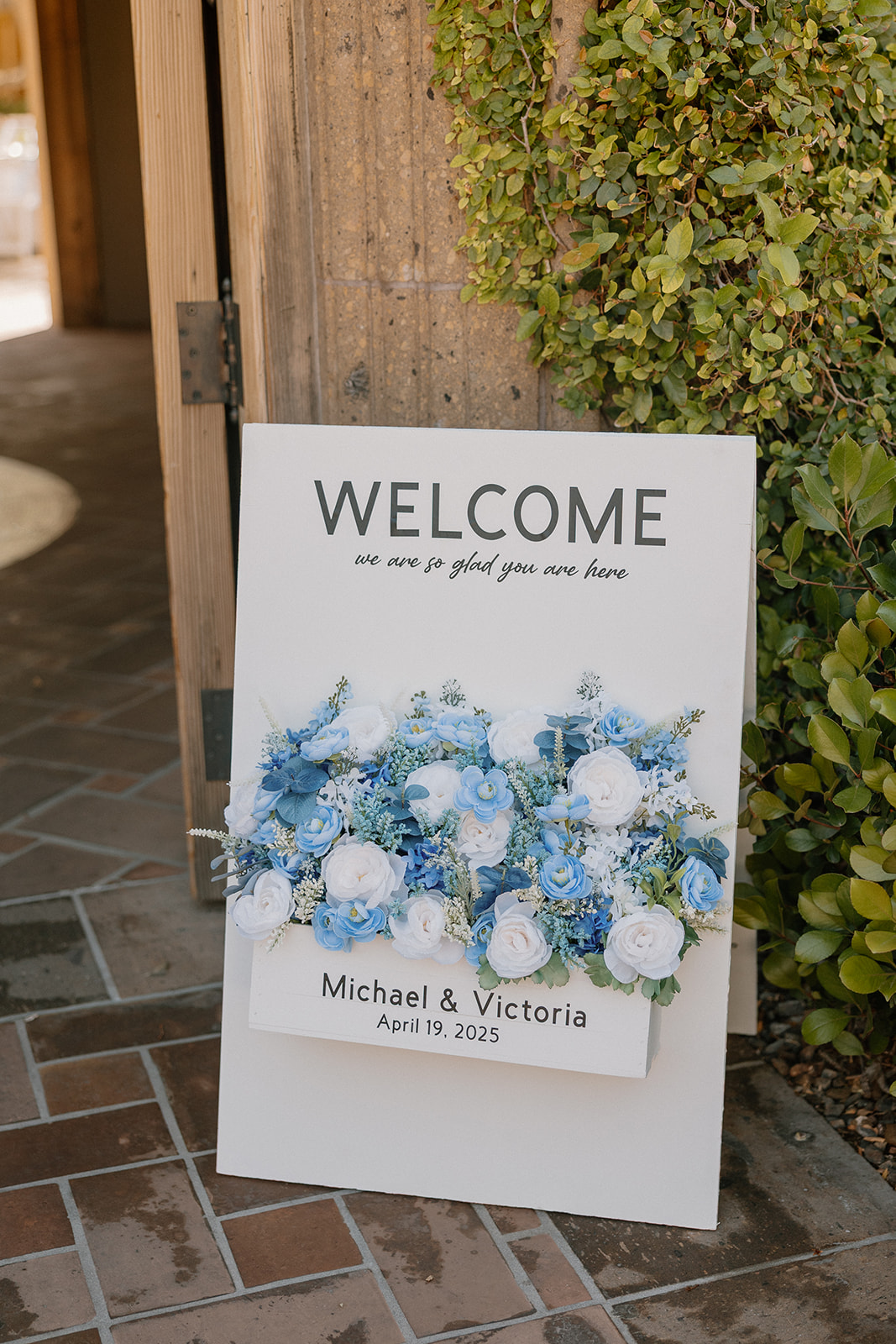 A welcome sign for Michael and Victoria’s wedding on April 19, 2025, adorned with blue and white florals and placed near the venue entrance.
