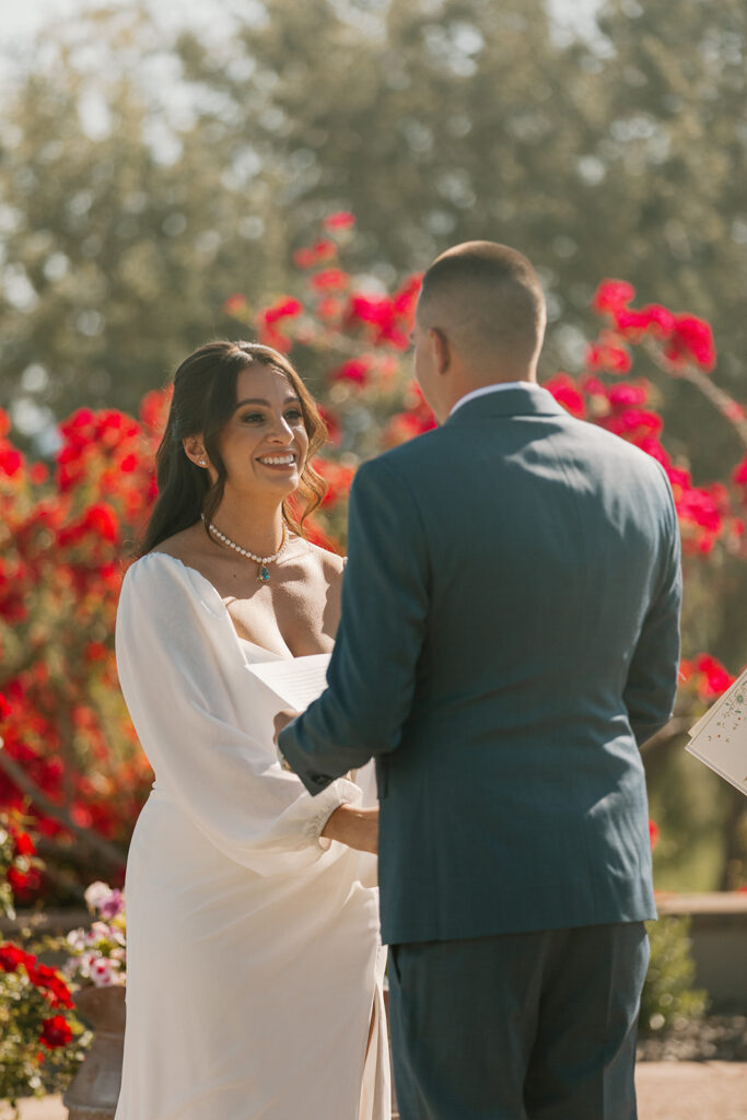 The bride beams at her groom during the ceremony, surrounded by red flowers and golden light.