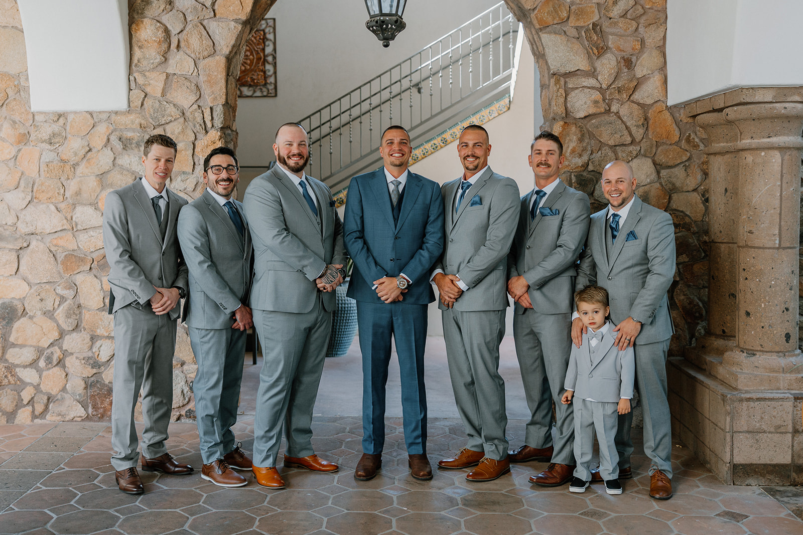 The groom and his groomsmen pose under the stone arch at Blackstone Country Club, dressed in sleek gray suits with blue accents.